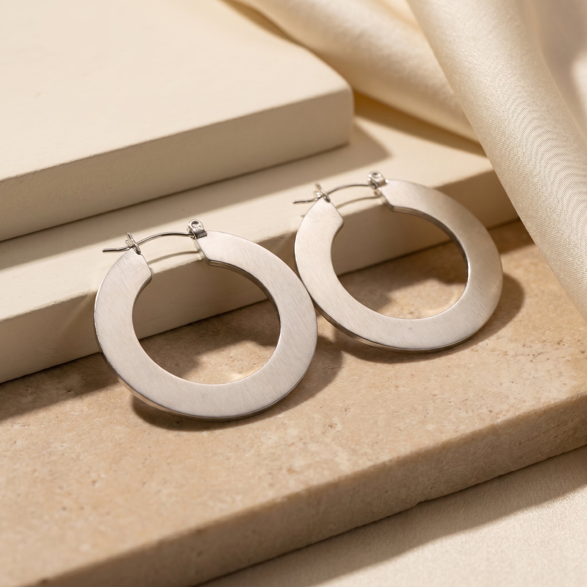 Matte silver PVD stainless steel hoop earrings resting on a textured stone and silk background.
