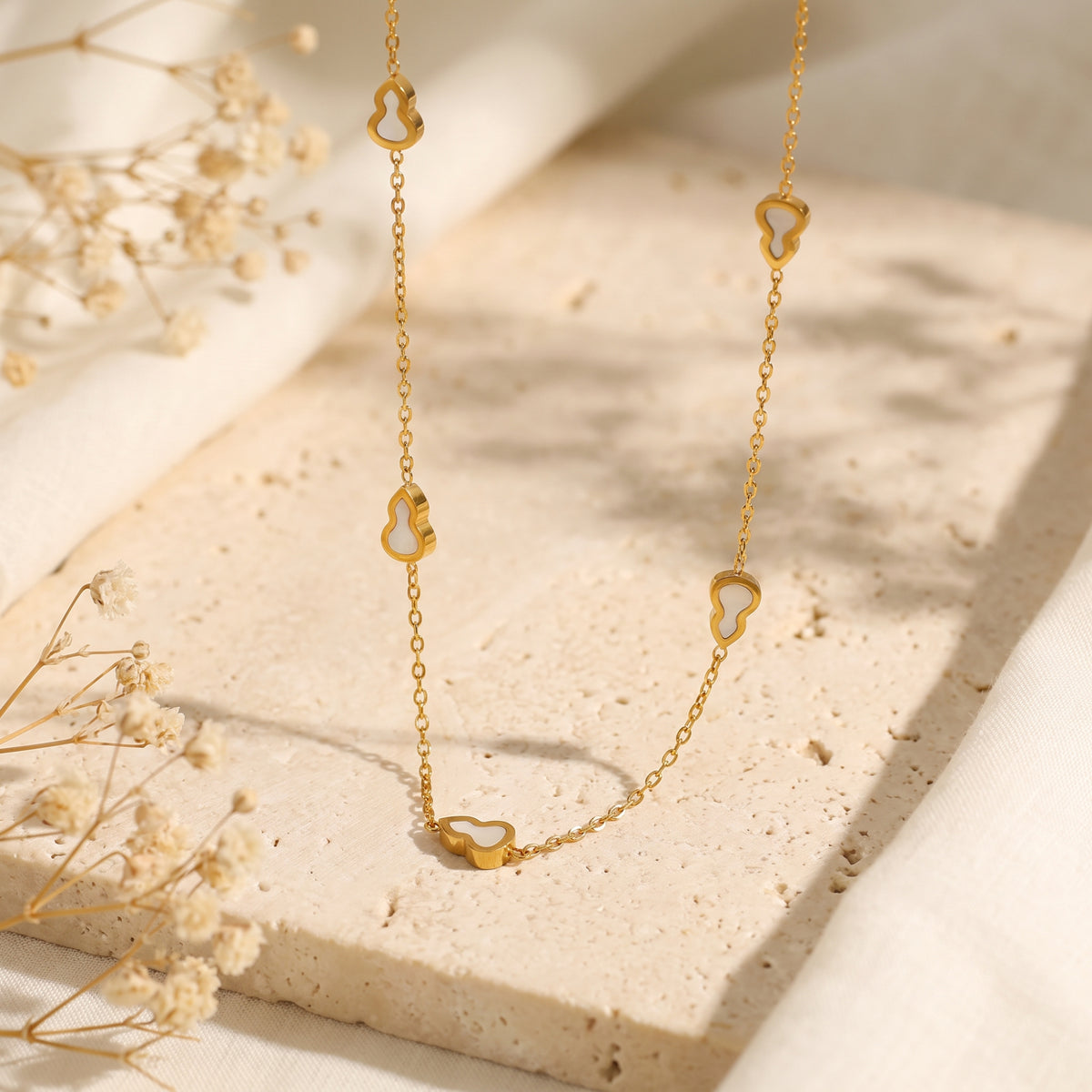 Gold-tone station necklace with white inlay gourd motifs arranged on a beige textured stone slab beside dried flowers.