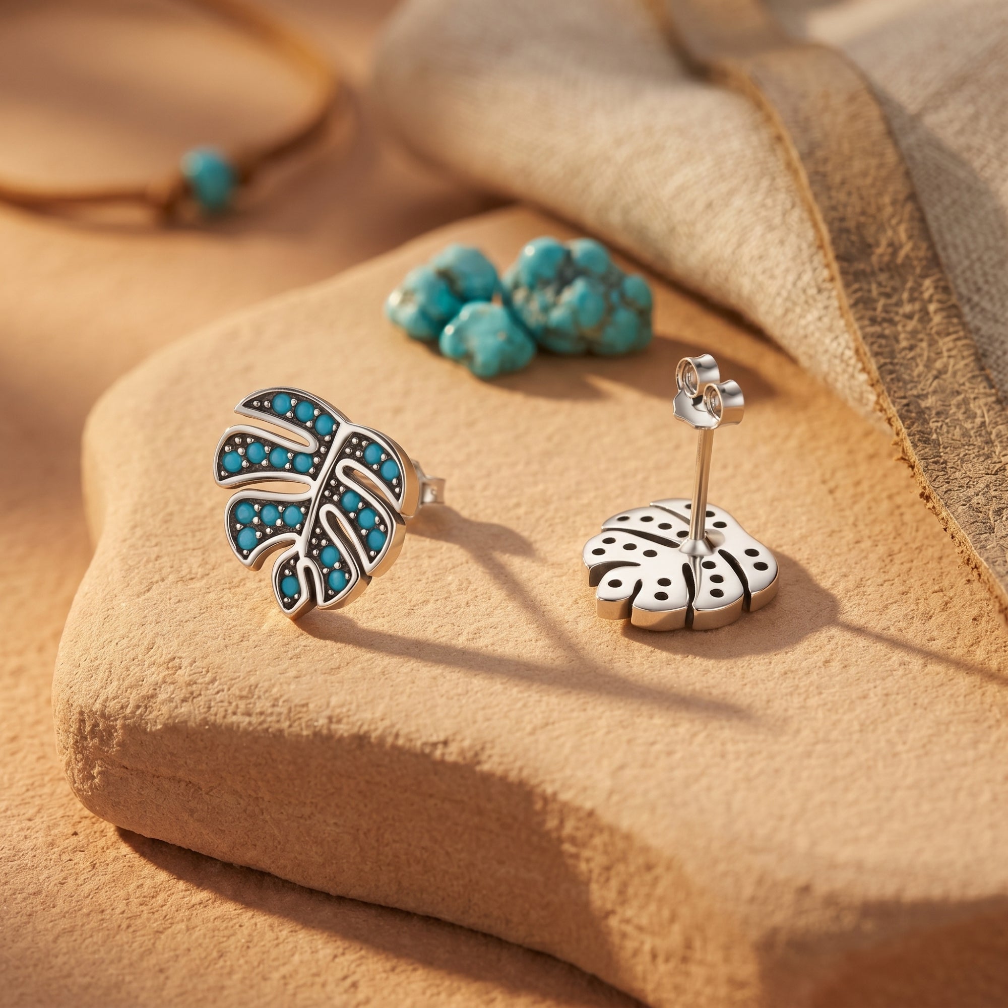 Front and back view of silver monstera leaf earrings with turquoise cabochons on a stone surface.