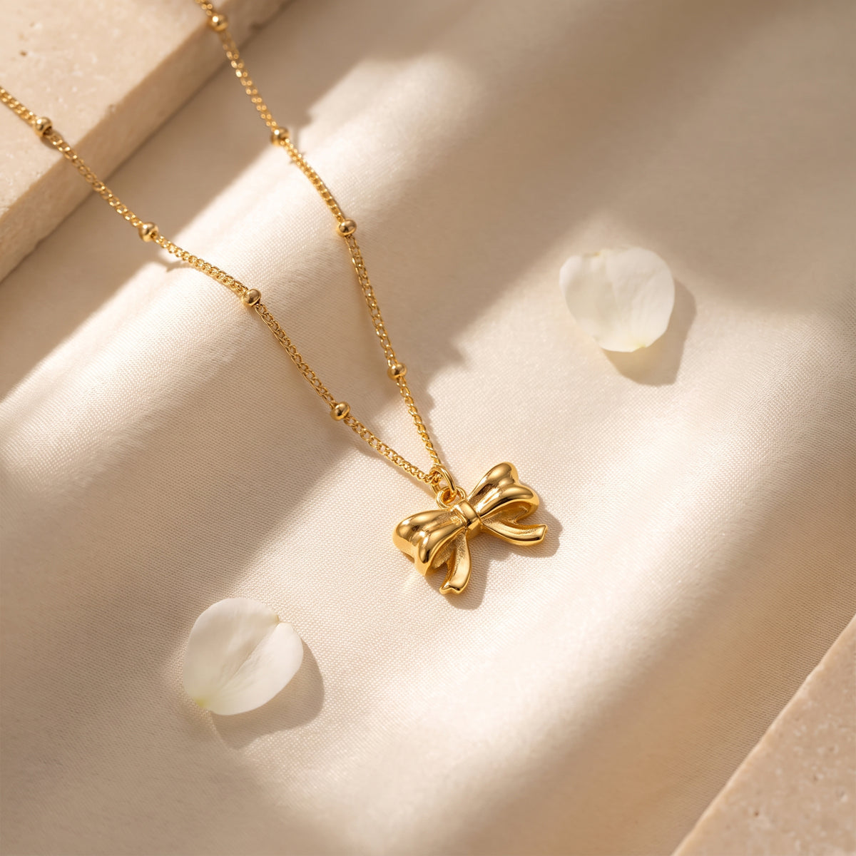 Close view of the gold-tone ribbon bow pendant on cream fabric beside scattered white flower petals.