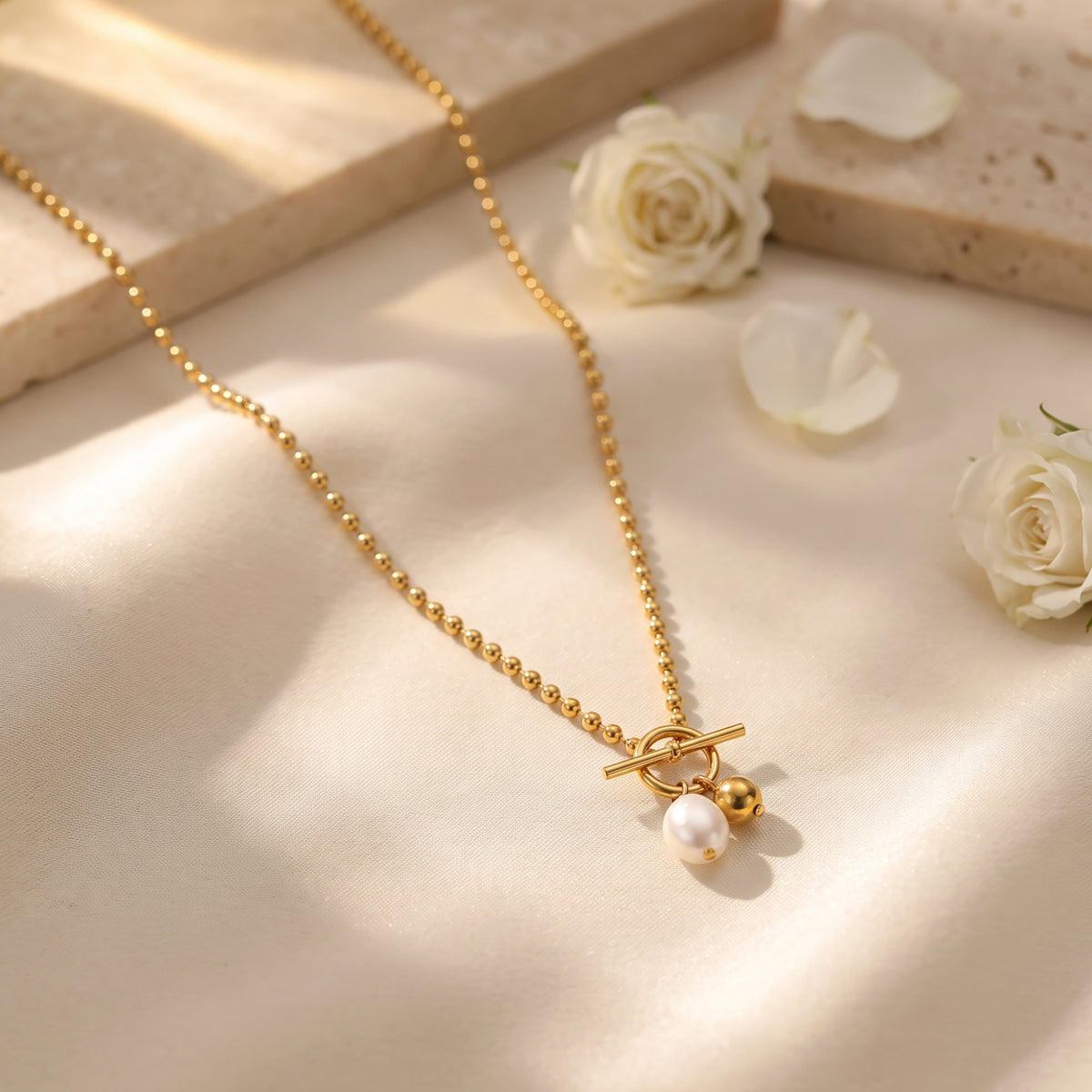 Gold-tone toggle necklace with pearl and sphere charms arranged on ivory fabric among white roses and stone blocks.