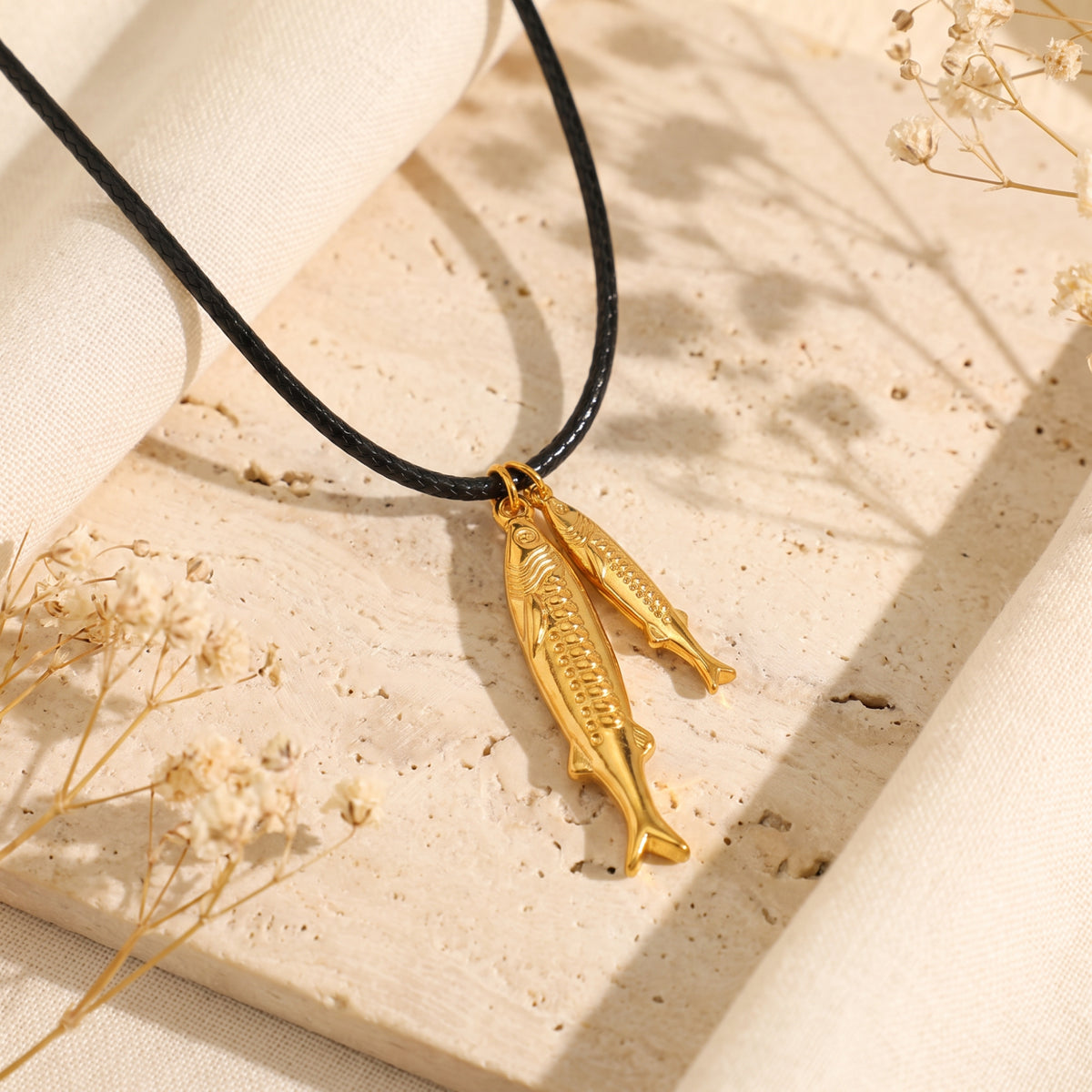 Angled view of the gold-tone double fish pendant on a black cord over a light textured stone surface.