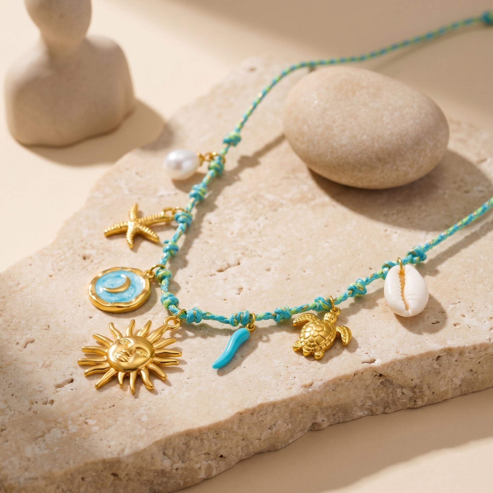 Close-up of gold sun, moon, and sea turtle charms on a turquoise woven cord necklace, resting on a stone slab.