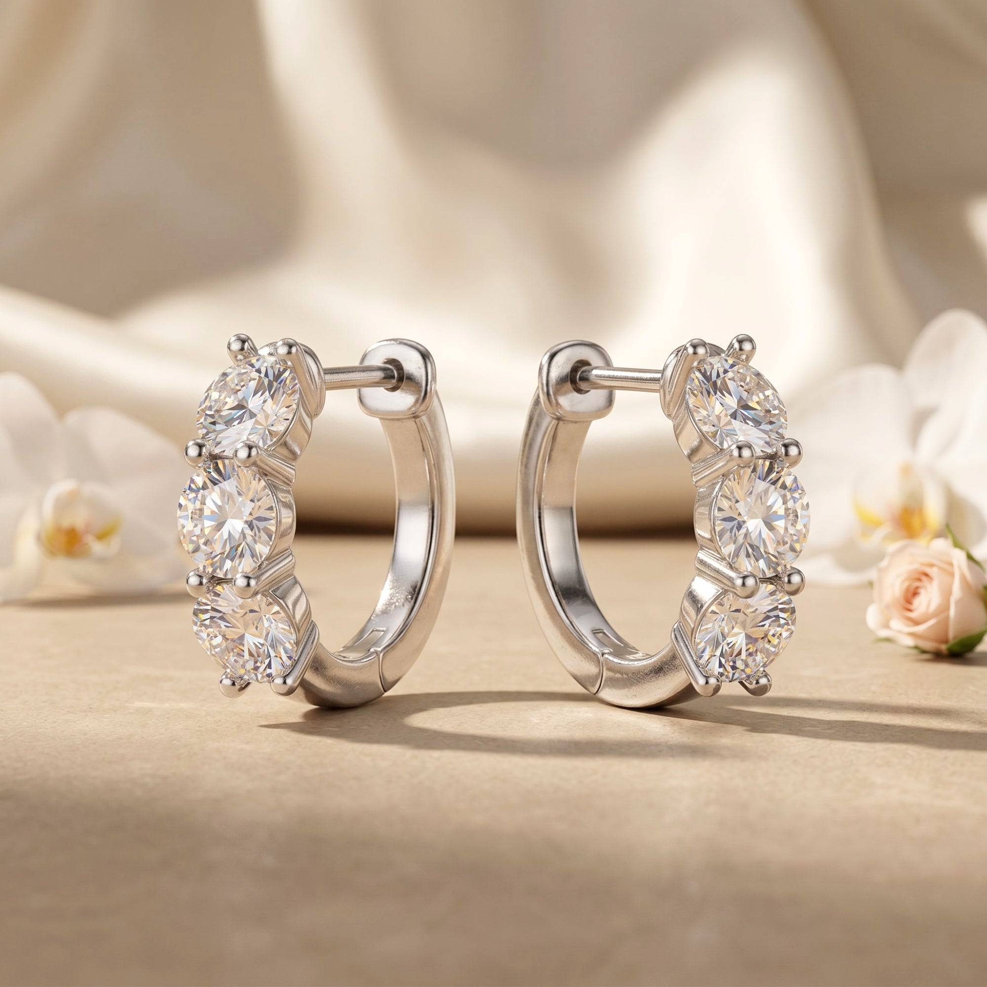 Silver moissanite hoop earrings with three round-cut stones on a neutral fabric background.