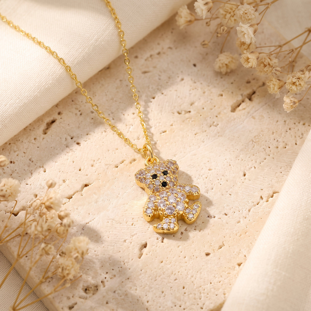 Angled view of a gold-tone crystal teddy bear pendant resting flat on a beige stone surface.