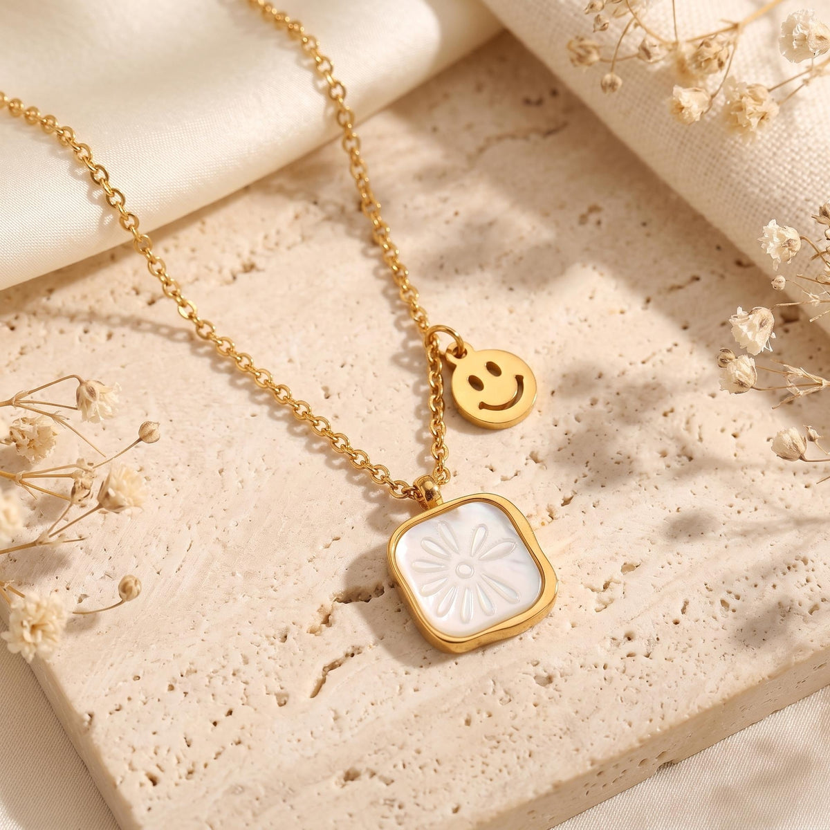 Gold-tone necklace with a white daisy pendant and smiley-face charm resting on a textured beige stone slab.