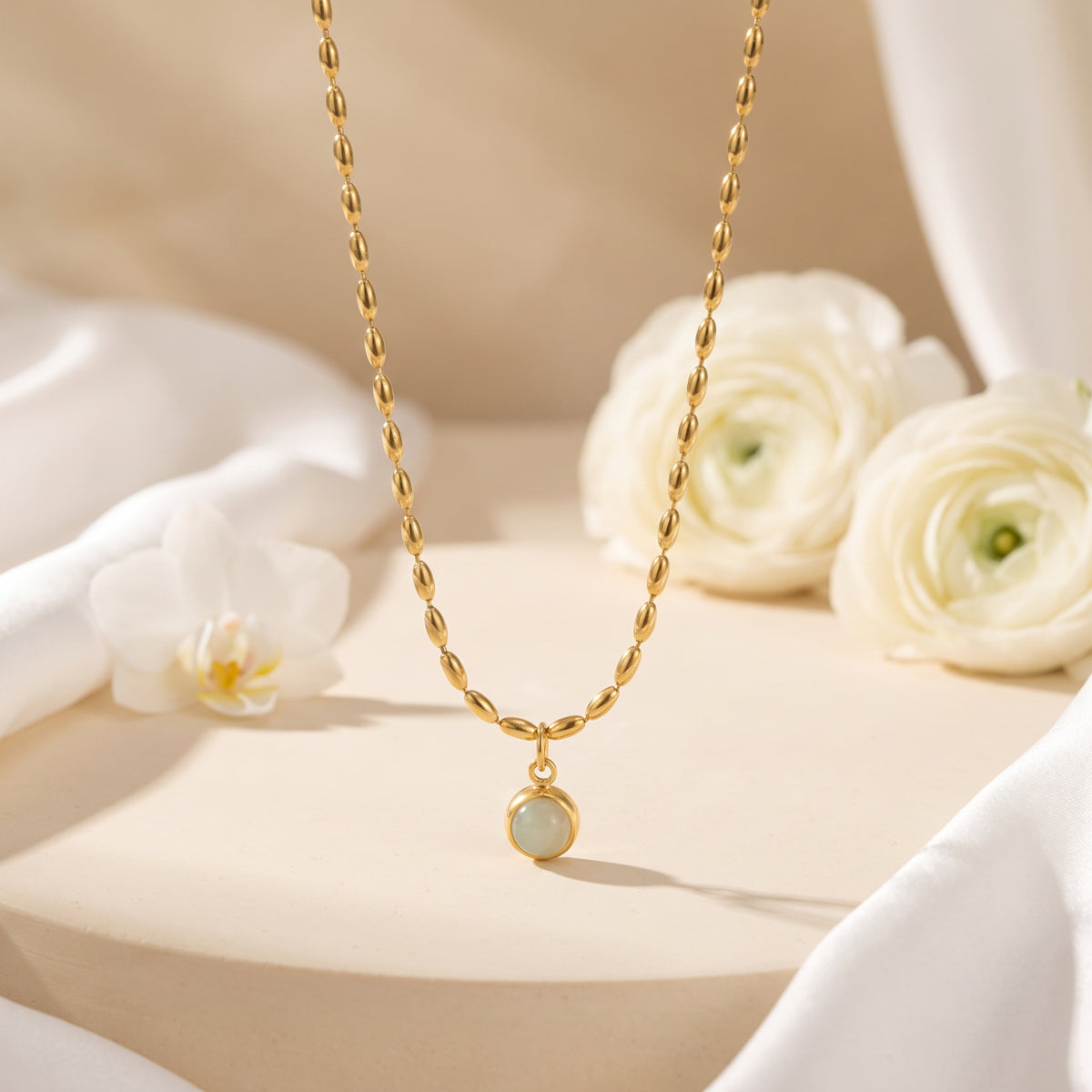 Gold-tone oval-bead pendant necklace displayed above a beige pedestal with white orchid and ranunculus flowers.