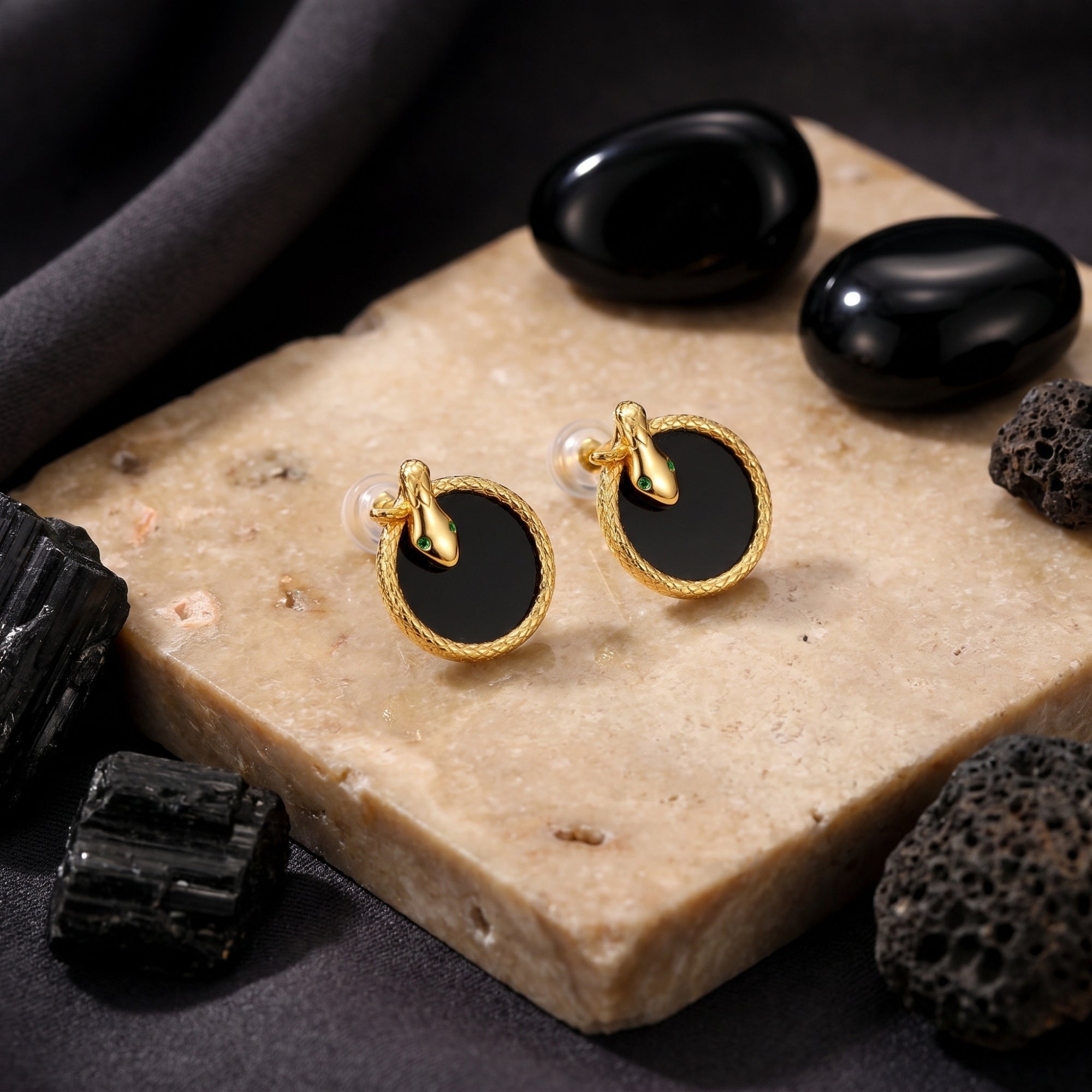 18K gold plated Serpens onyx earrings with snake detail displayed on a textured stone and volcanic rock surface.