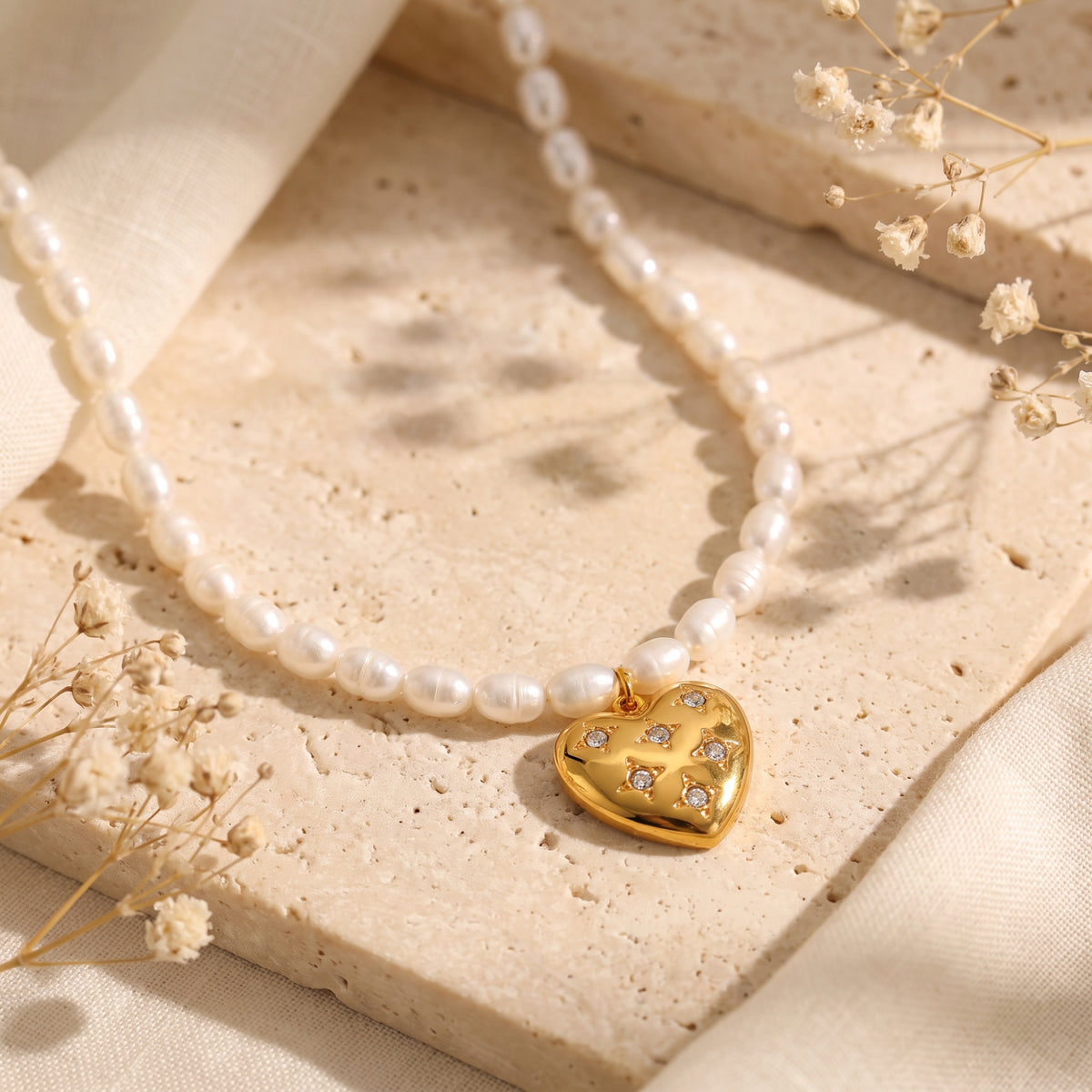 White pearl necklace curved around a gold-tone puffed heart pendant on a stone tile.