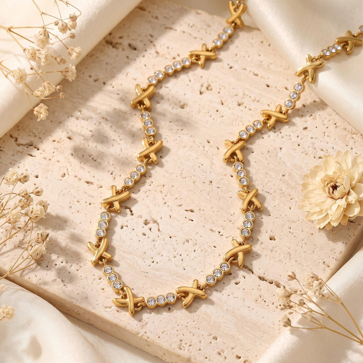 Flat lay of the gold-tone X station necklace on a textured beige stone slab with dried accents.