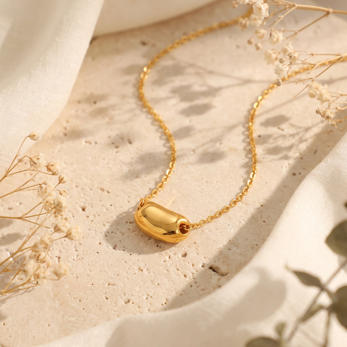 Gold-toned pod pendant necklace resting on a textured stone board with cream fabric and dried flowers.