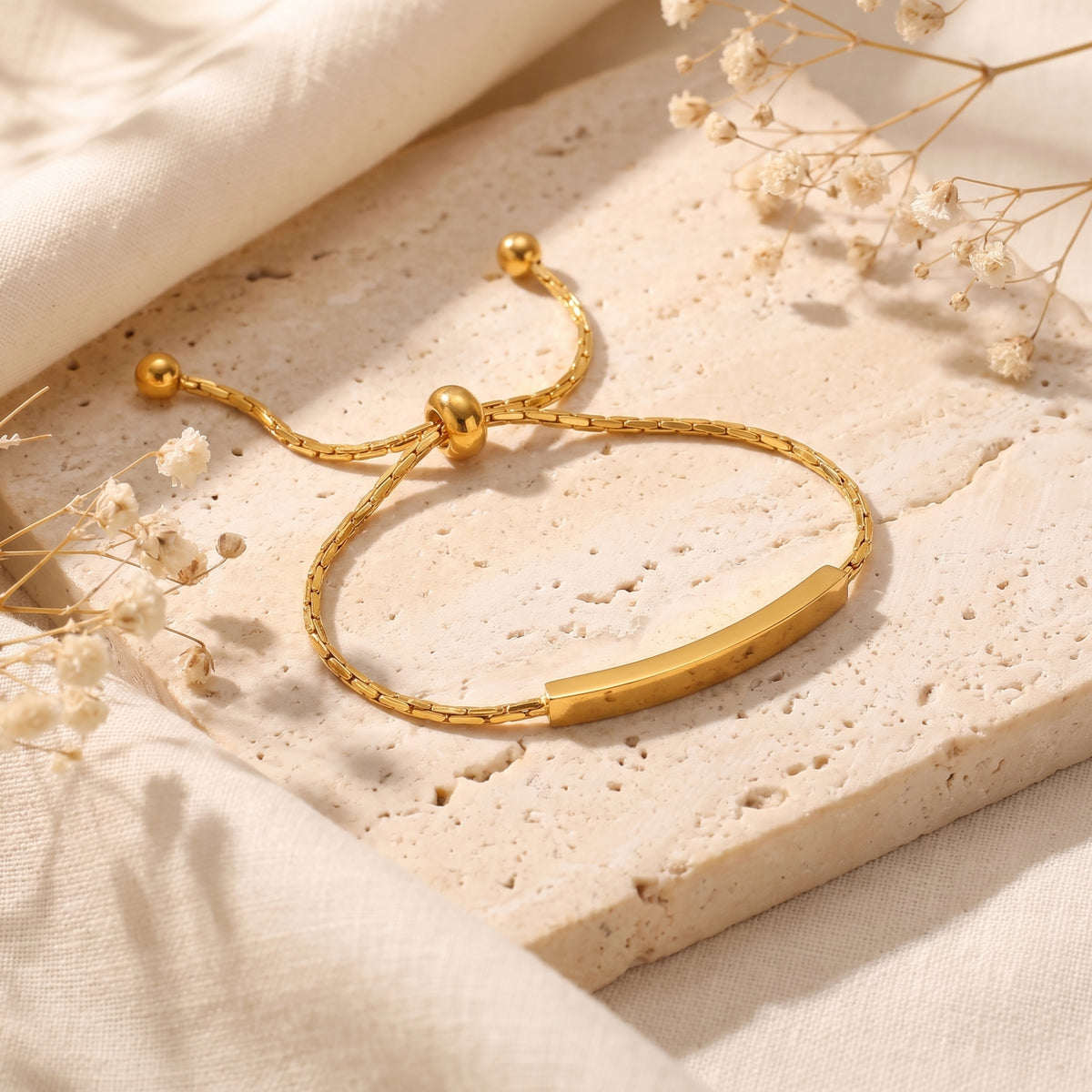 Angled view of a gold-tone curved bar slider bracelet on a pale stone slab beside dried flowers.