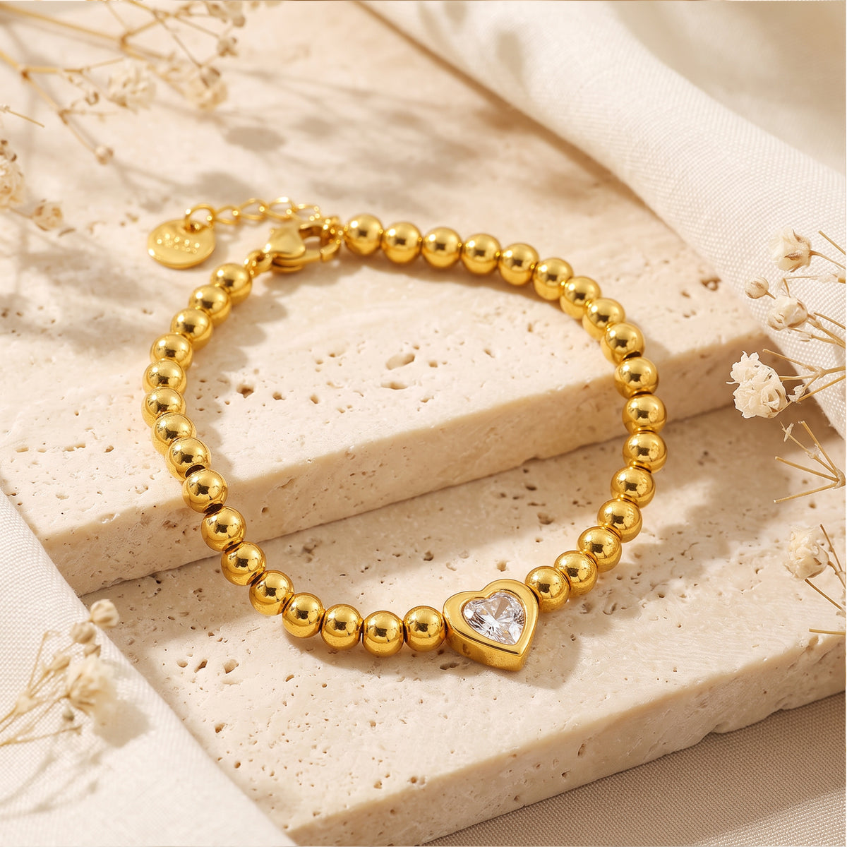 Gold-tone beaded bracelet with a heart-shaped clear stone arranged on a porous beige stone slab with dried flowers.