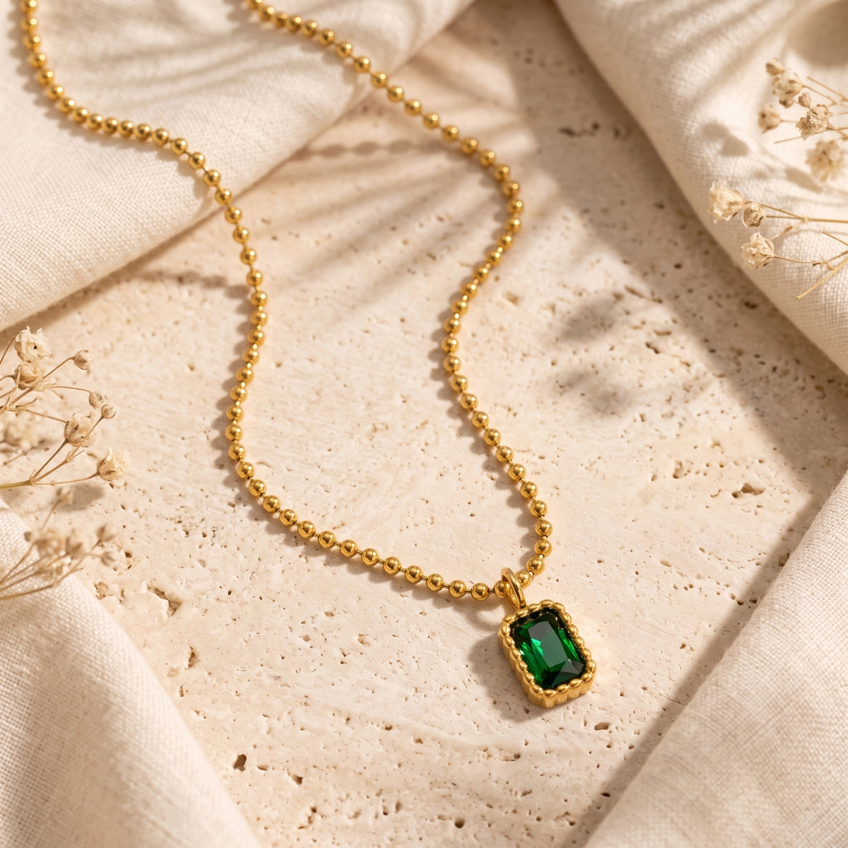 Flat lay view of the green emerald-cut pendant necklace and gold-tone ball chain on textured stone.