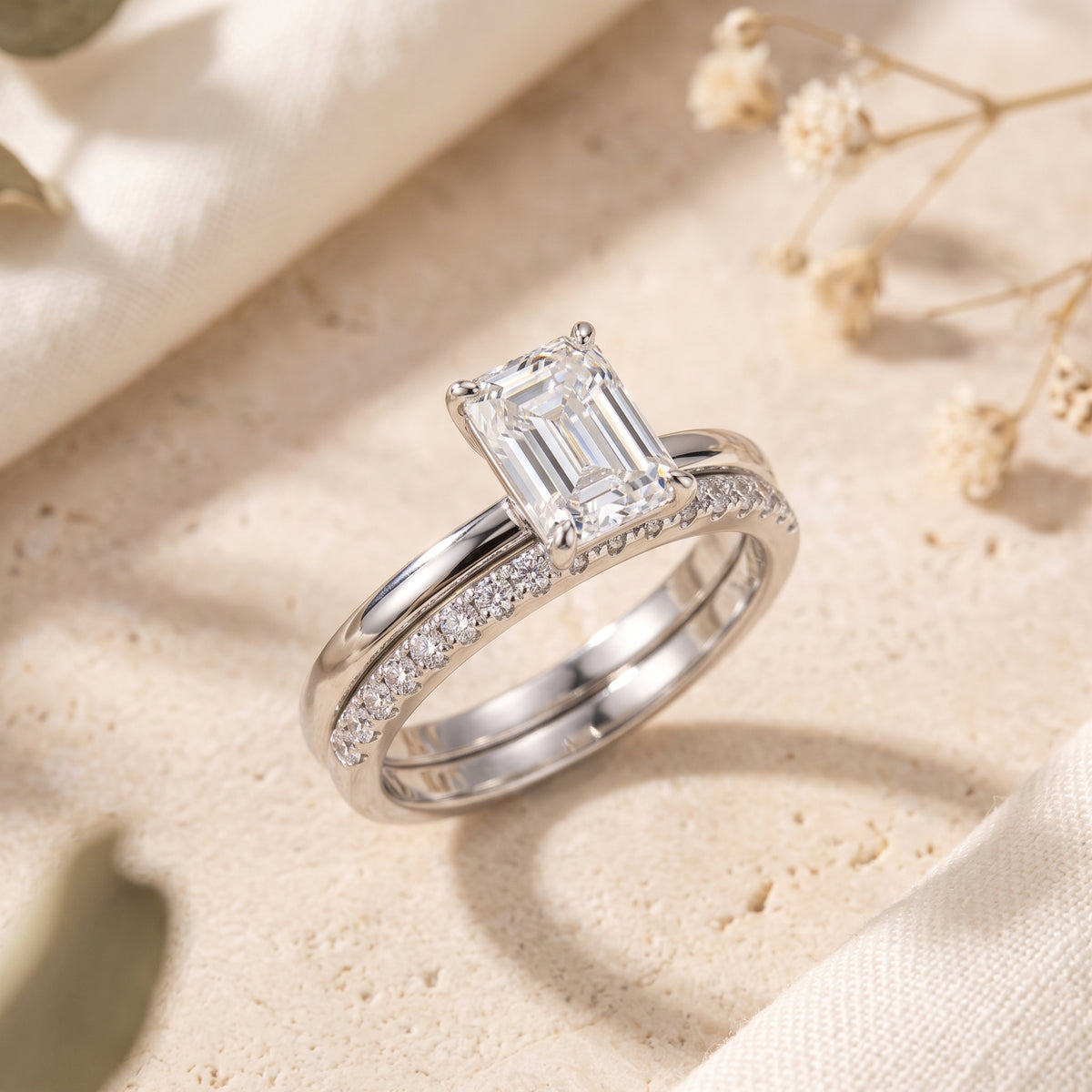 Two silver rings overlapping on a stone surface, showing an emerald-cut center stone and a matching pavé band.