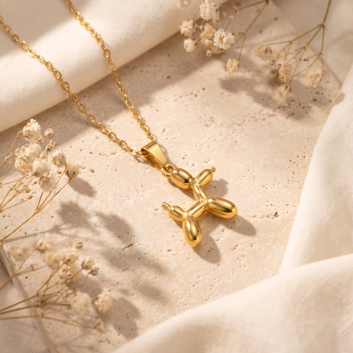 Gold-tone balloon dog pendant necklace lying on a textured beige stone-like slab with dried flowers.