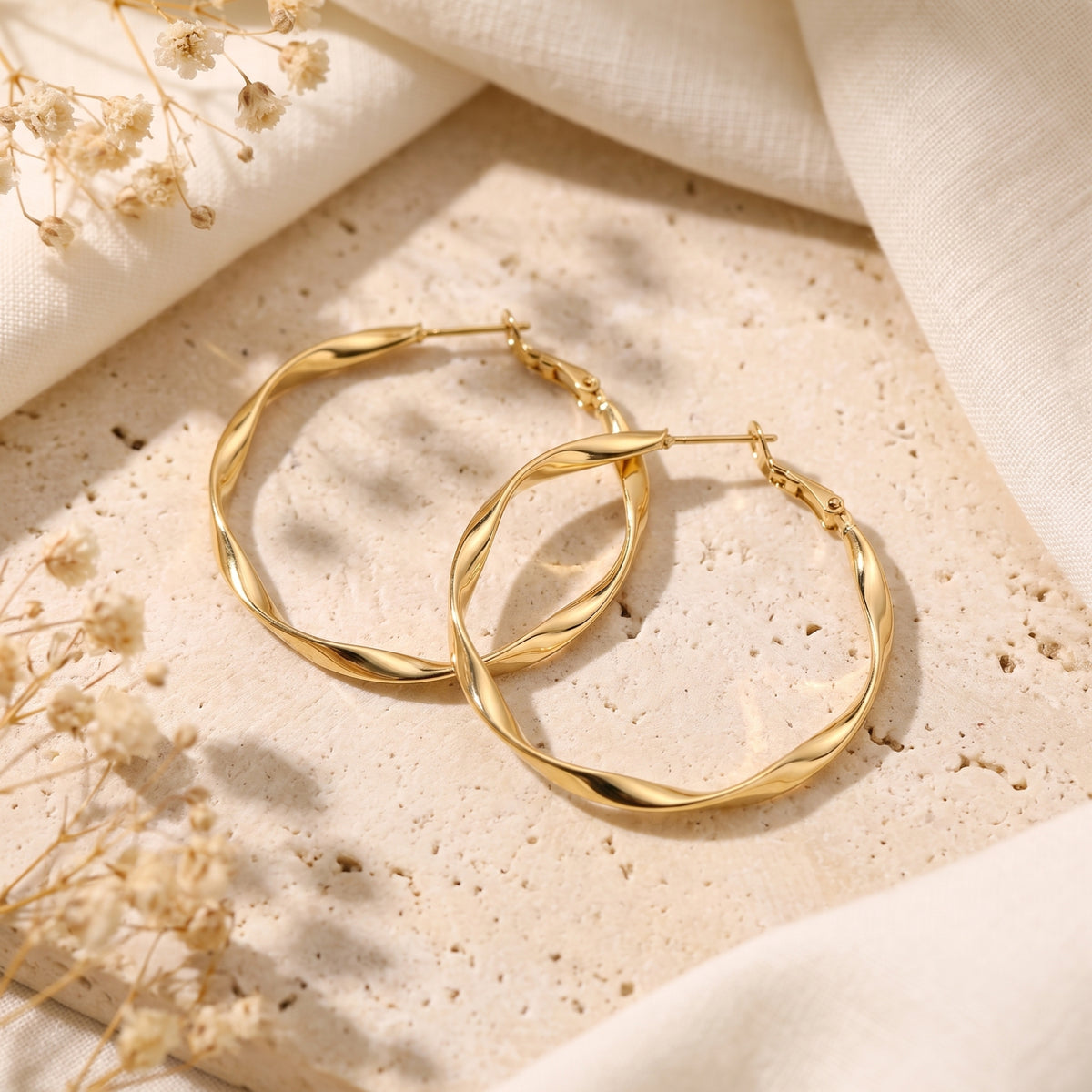 Overhead view of both gold-finish twisted oval hoop earrings resting on a light stone plaque.