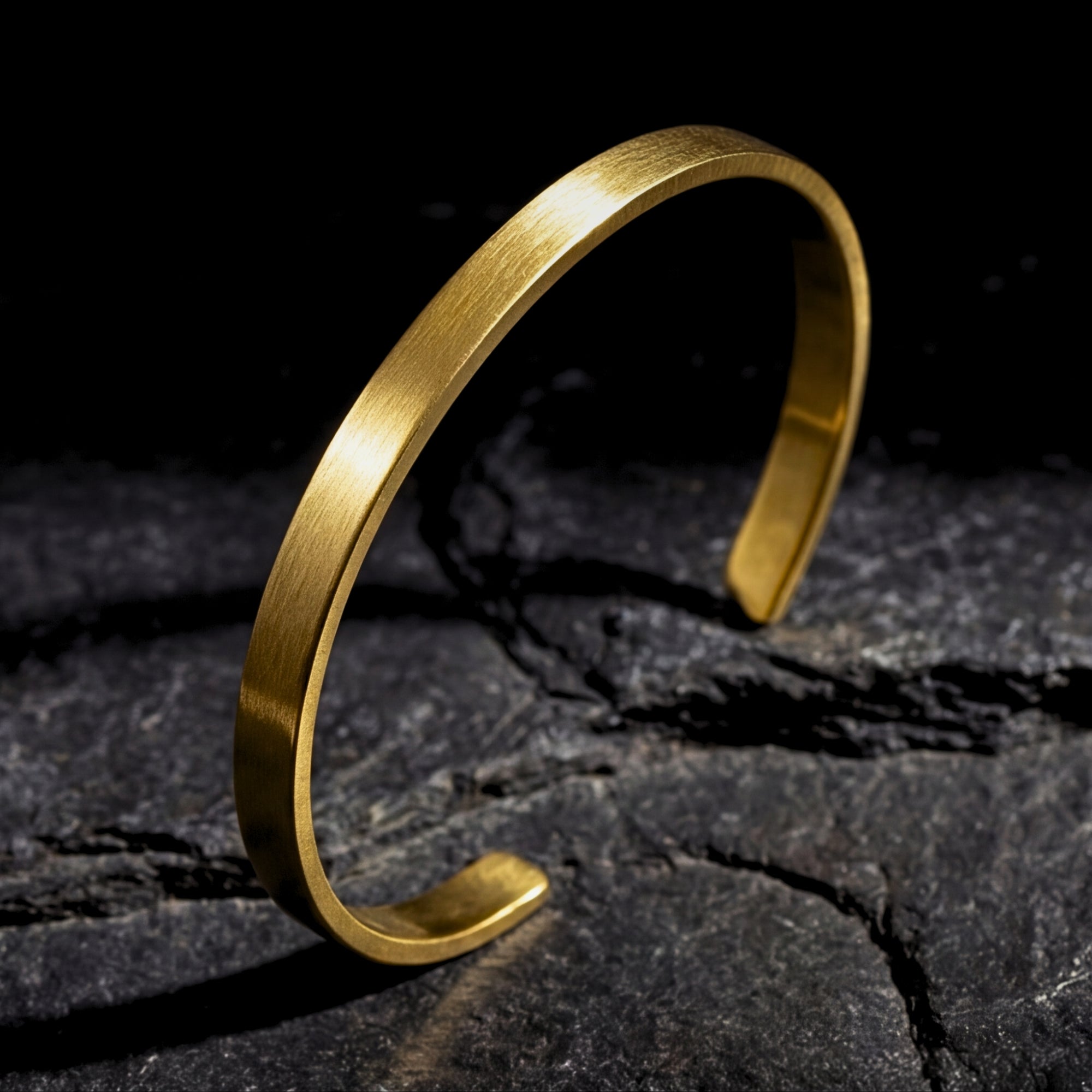Brushed gold stainless steel Meridian Cuff bracelet standing upright on a dark, textured rock surface.