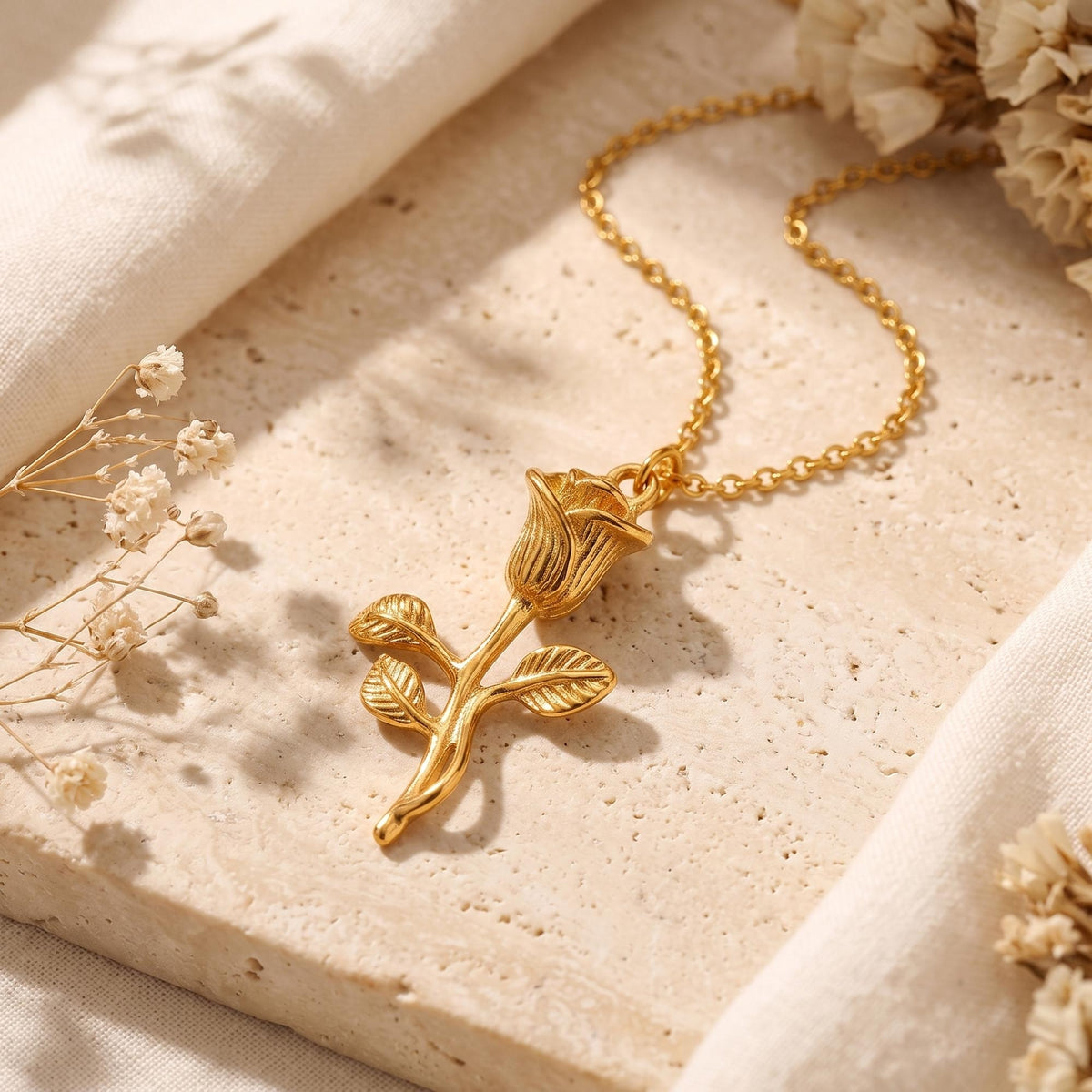 Close-up of a gold-tone rose pendant with a stem and leaves on a beige stone surface.