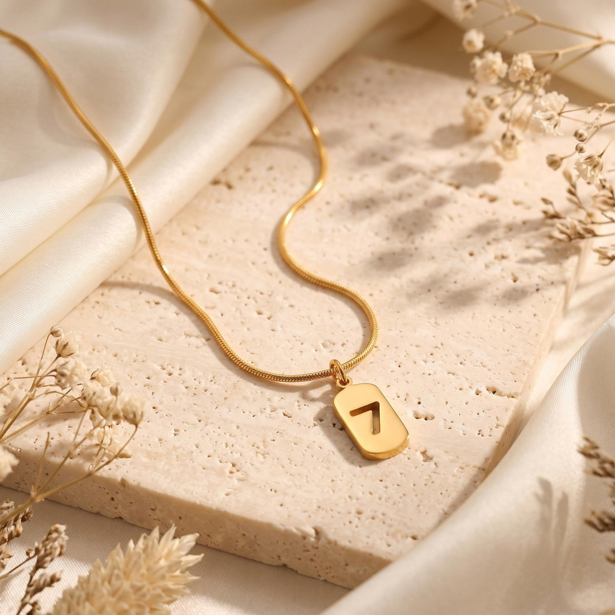 Gold-tone number 7 cutout pendant necklace displayed on a stone slab with silk fabric and dried flowers.