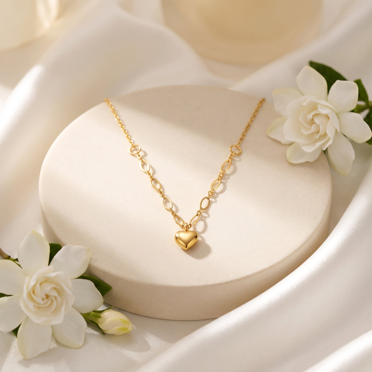 Gold-tone puffy heart necklace displayed on a round cream pedestal surrounded by white flowers.
