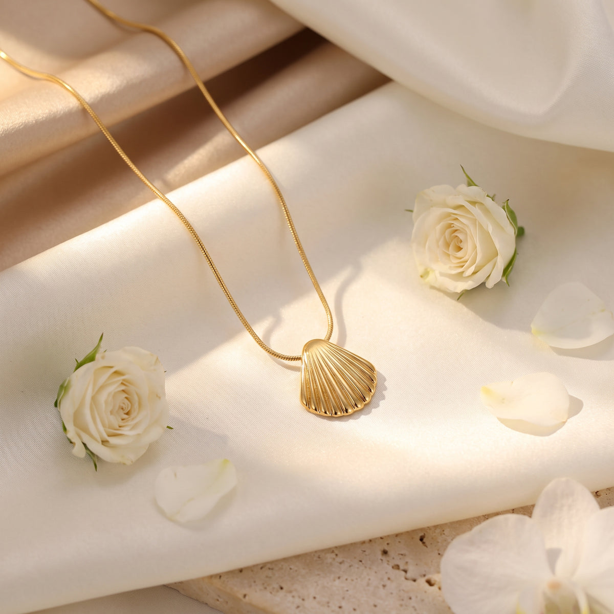 Gold scallop shell pendant necklace arranged on cream satin with white roses and scattered petals.