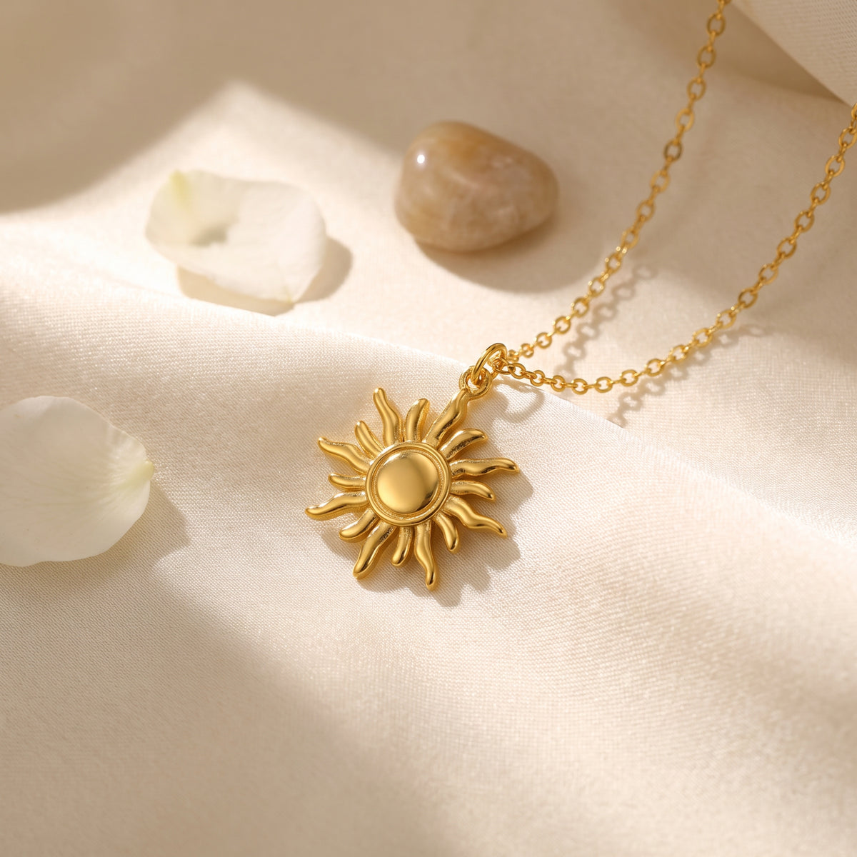 Gold sunburst pendant necklace lying on cream silk beside white petals and a polished stone.