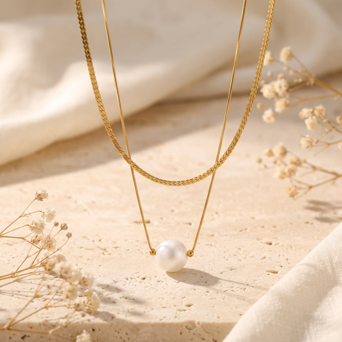 Gold-tone layered necklace arranged on a pale stone surface, with a white pearl pendant and dried flowers in the background.