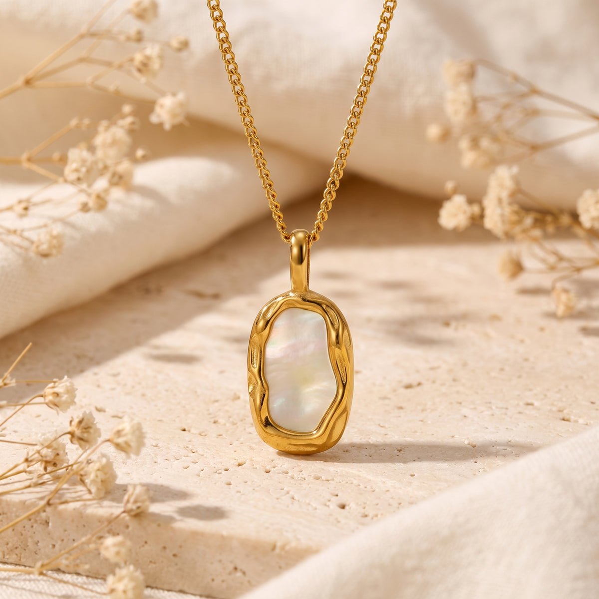 Gold-tone shell pendant necklace displayed upright on a beige stone surface with dried flowers.
