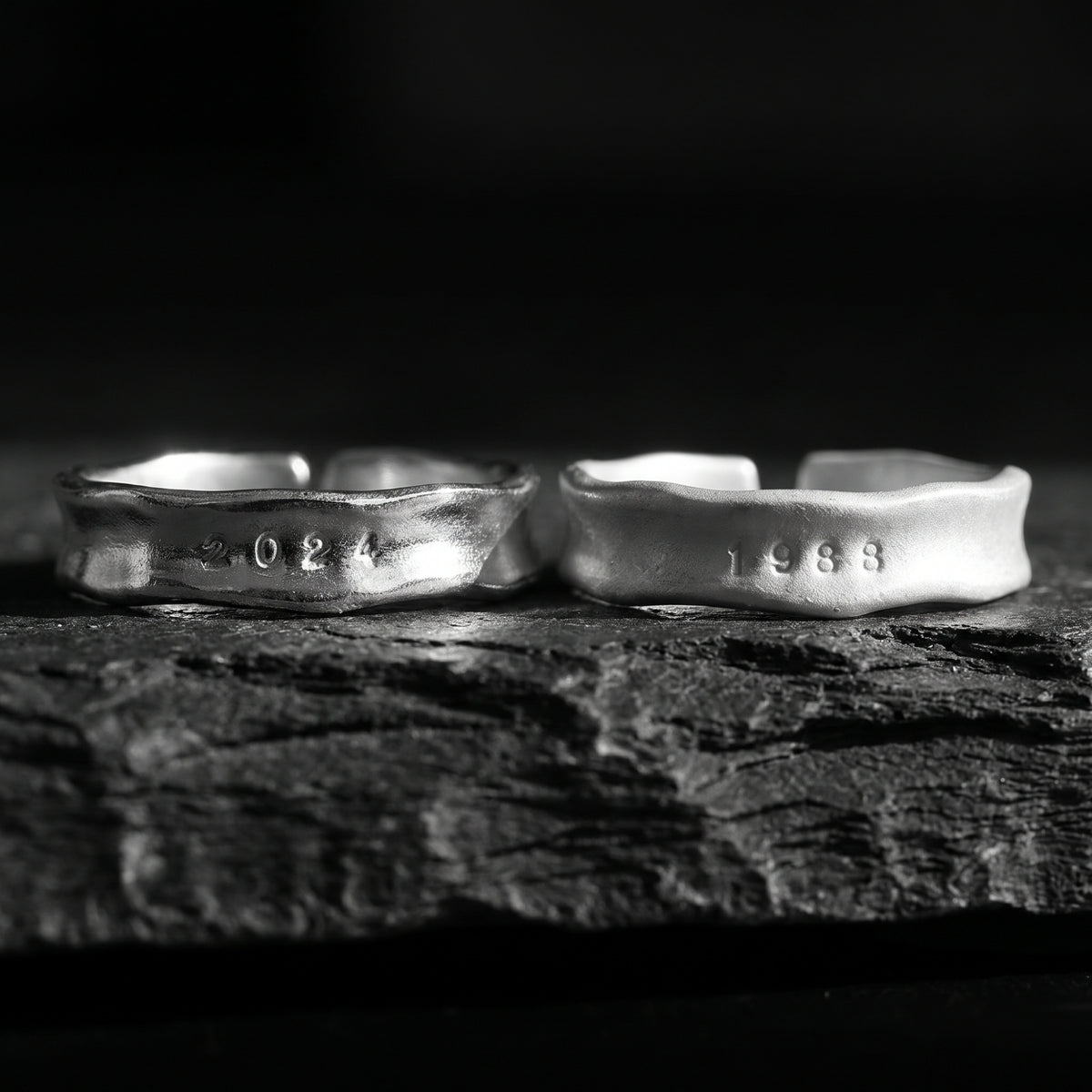 Two textured silver open-band rings stamped 2024 and 1988 displayed side by side on a rough stone surface