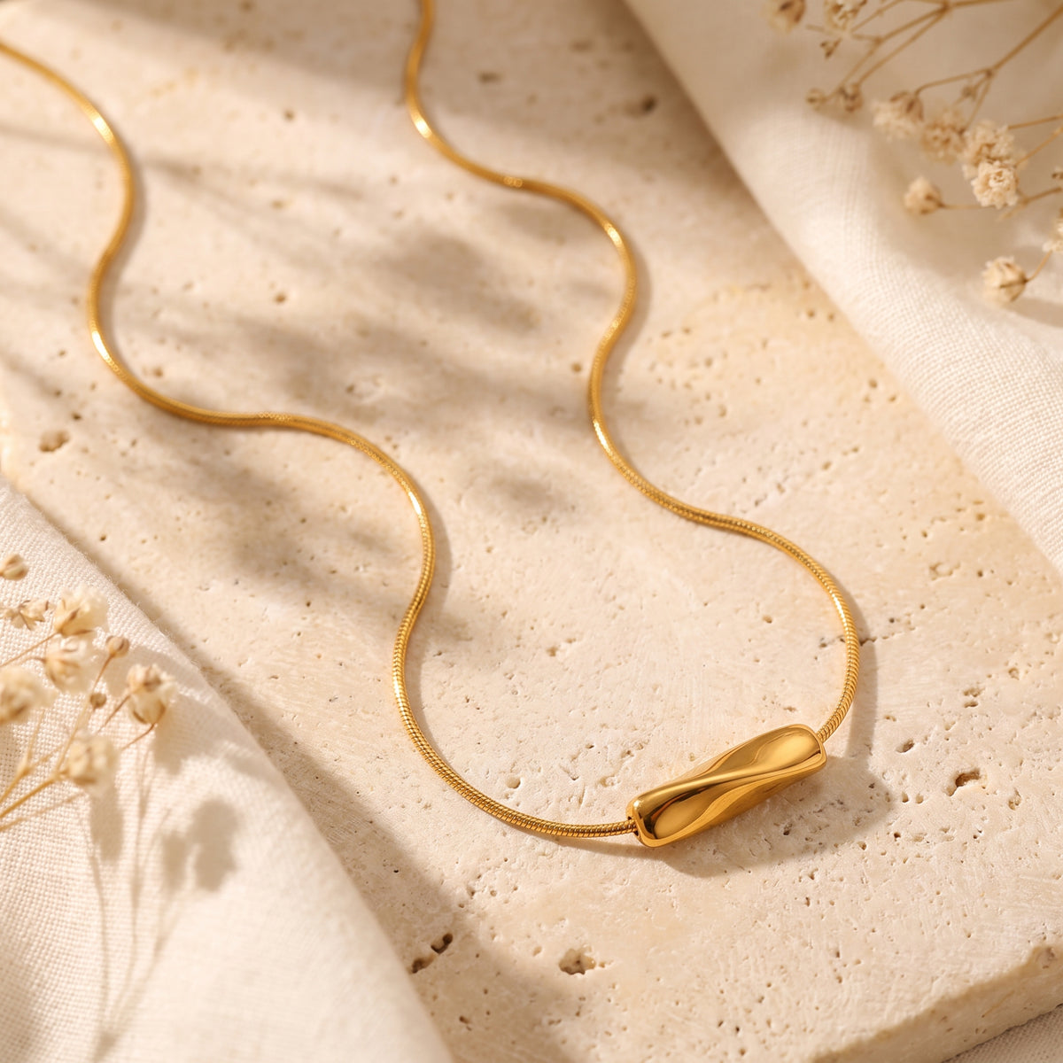 Flat lay of a gold-tone wave slide necklace on textured beige stone with dried flowers.