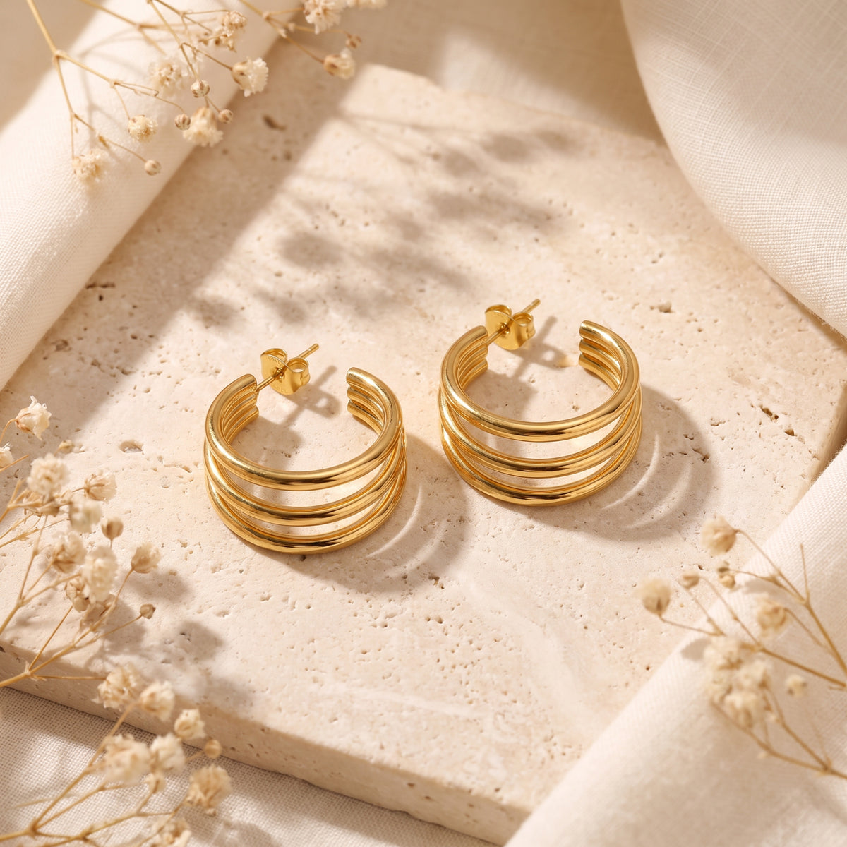 Top-down view of a pair of gold-color triple-band C-hoop earrings laid flat on a beige stone tile.
