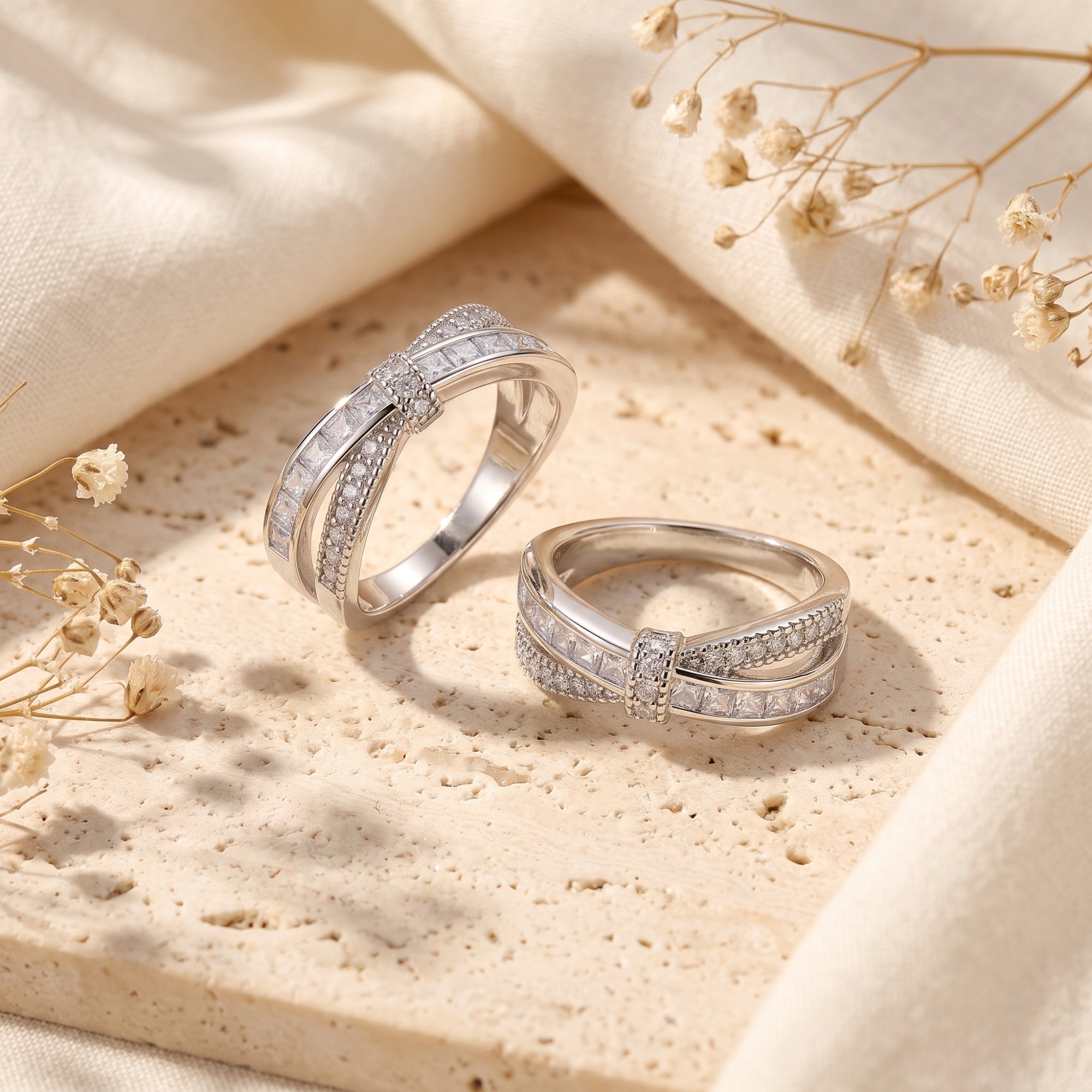 Two Charlotte sterling silver ribbon rings with overlapping CZ bands displayed on a stone surface with flowers.