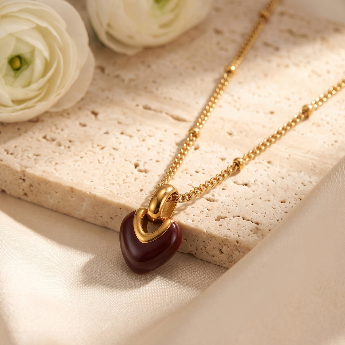 Angled detail shot of the gold-tone beaded necklace and deep red heart pendant resting on a stone surface.