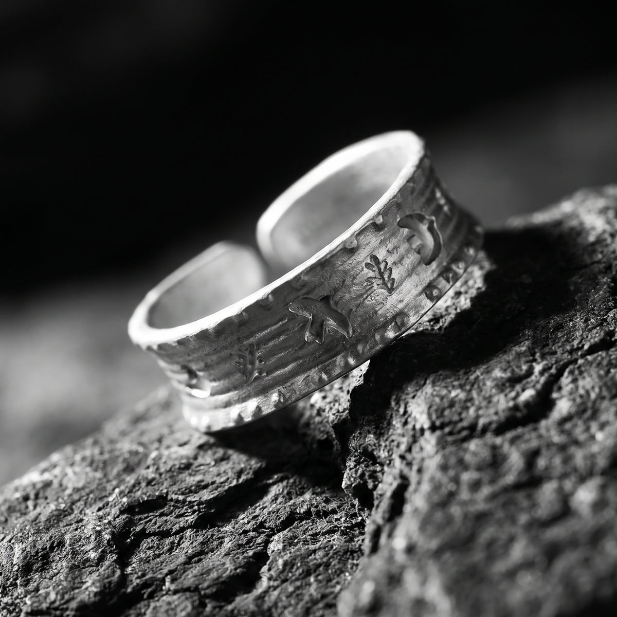 Angled close-up of a wide silver open-band ring with raised motifs and textured surface on dark rocks.