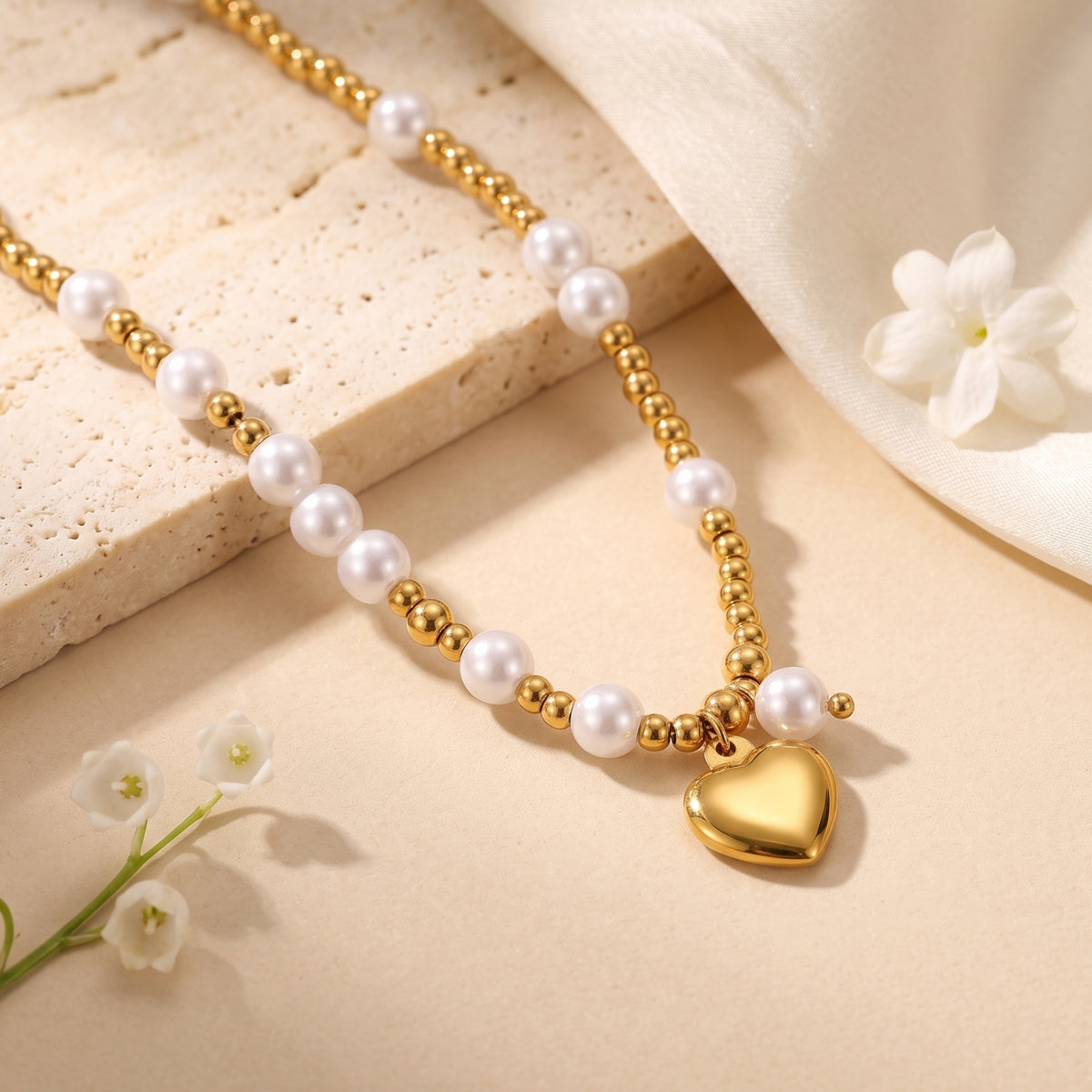 Gold-tone heart pendant necklace with white pearls laid on a beige stone slab beside white flowers and fabric.