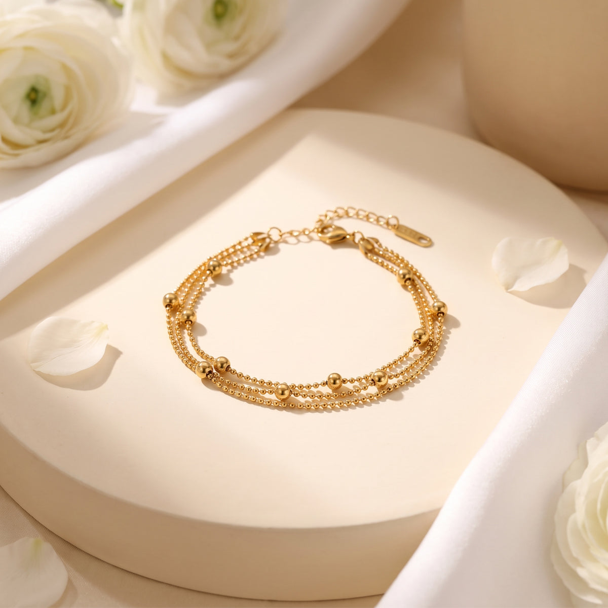 Gold-tone three-strand bead bracelet displayed on a round pedestal with white flowers and petals.