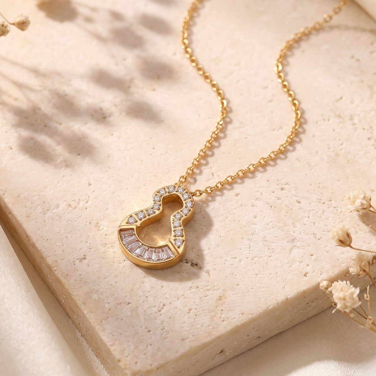 Angled shot of the gold-tone keyhole pendant and chain laid on a textured stone surface with dried flowers.