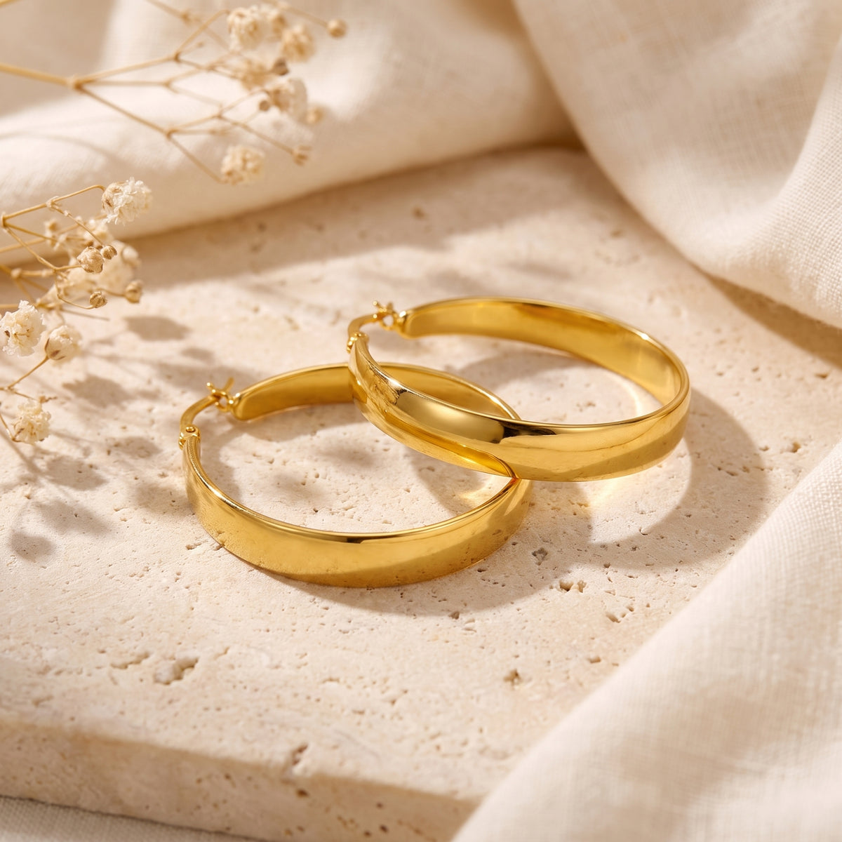 Pair of gold-tone tapered hoop earrings lying flat and overlapping on a neutral stone display.