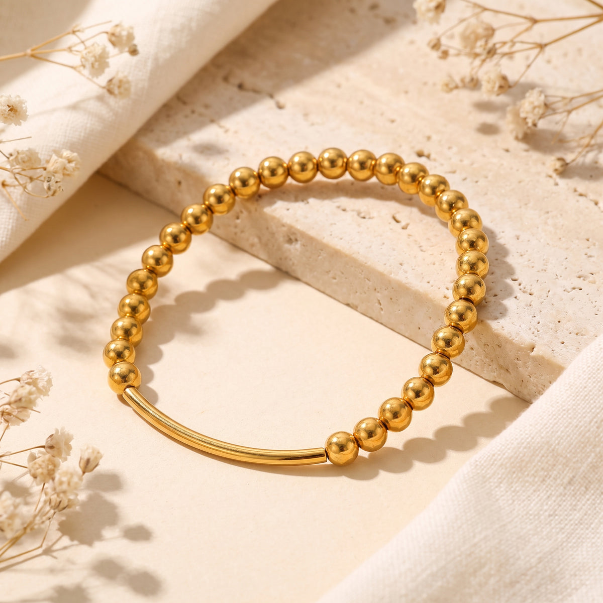 Gold-tone beaded bracelet with a curved bar centerpiece resting on a textured stone slab.