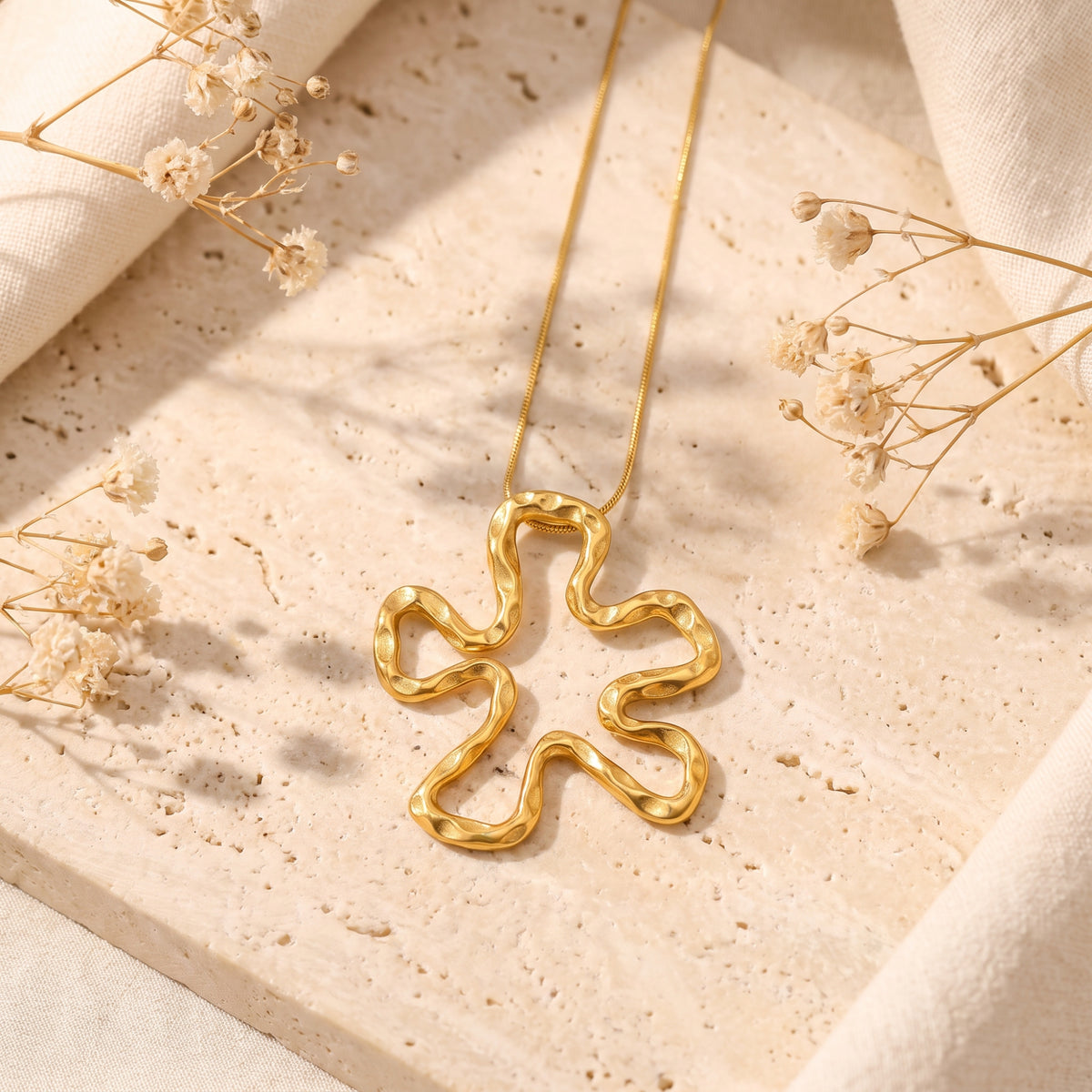 Top-down view of the gold-tone openwork wavy flower pendant lying on a beige textured stone slab among dried flowers.
