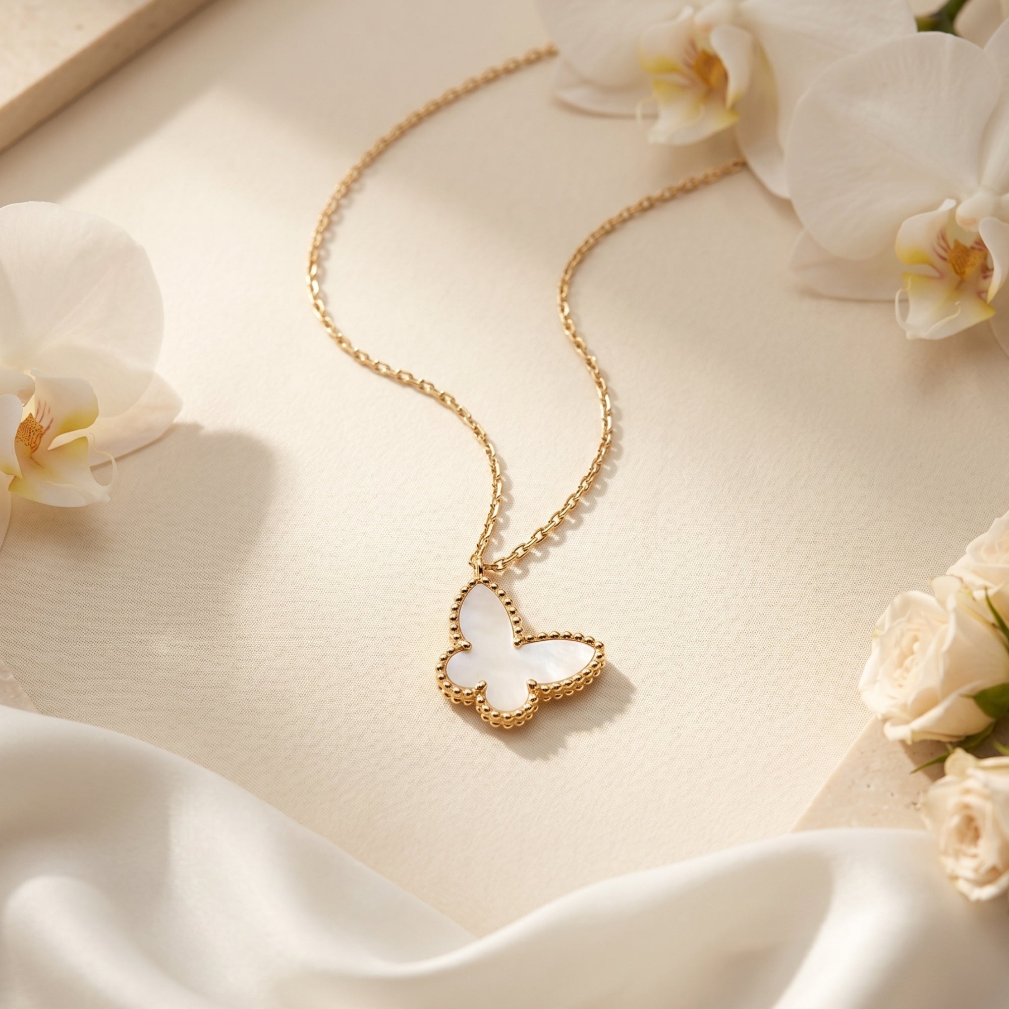 Yellow gold mother of pearl butterfly necklace displayed flat on a cream surface with white roses and silk.