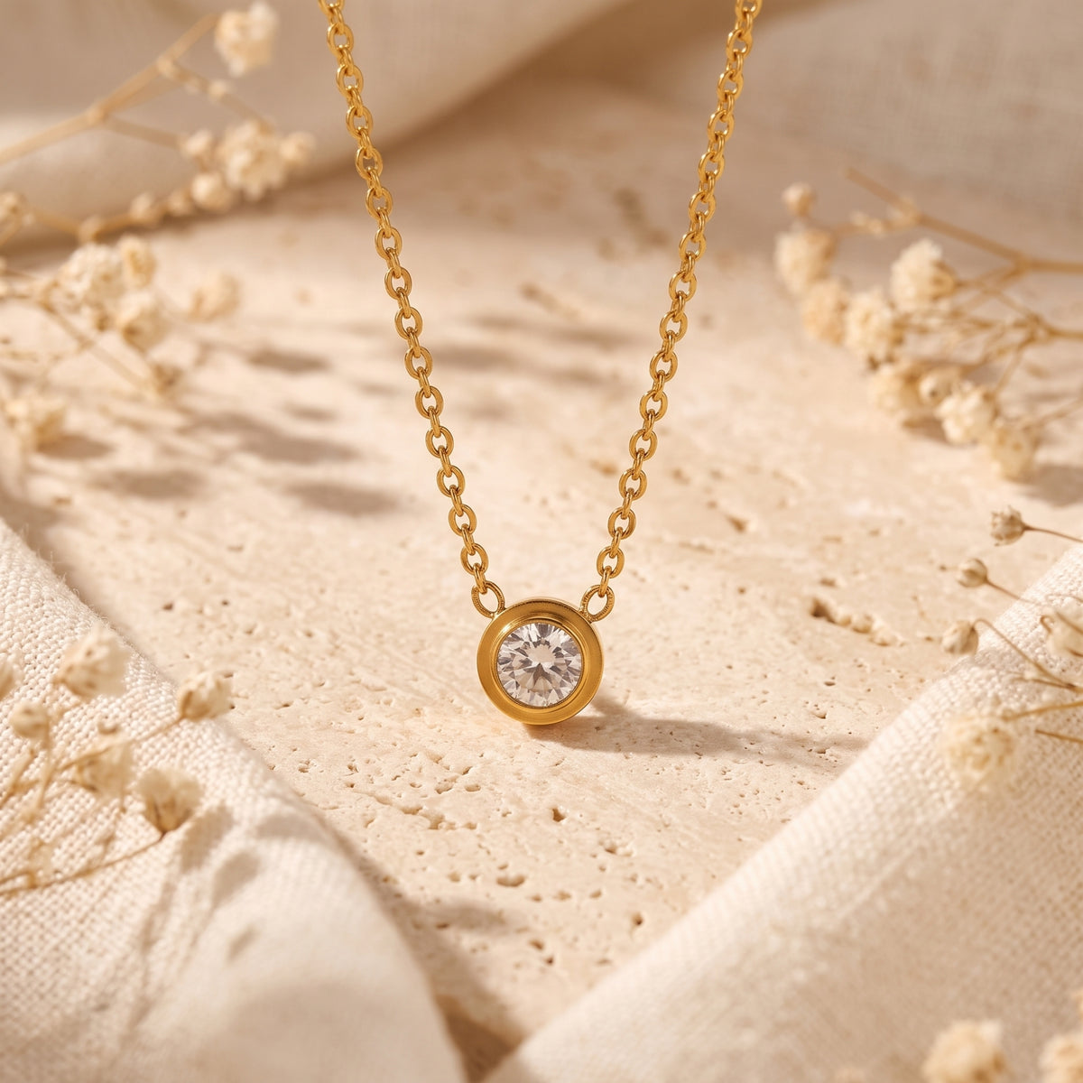 Gold-tone bezel solitaire necklace displayed on a pale textured stone surface with white fabric and dried flowers.