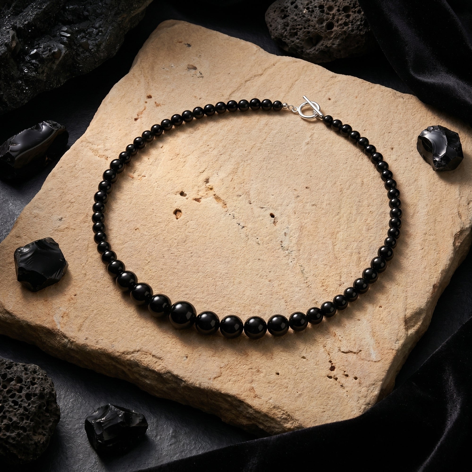 Angled view of a natural black onyx graduated bead necklace with silver clasp on a stone slab with raw black minerals.