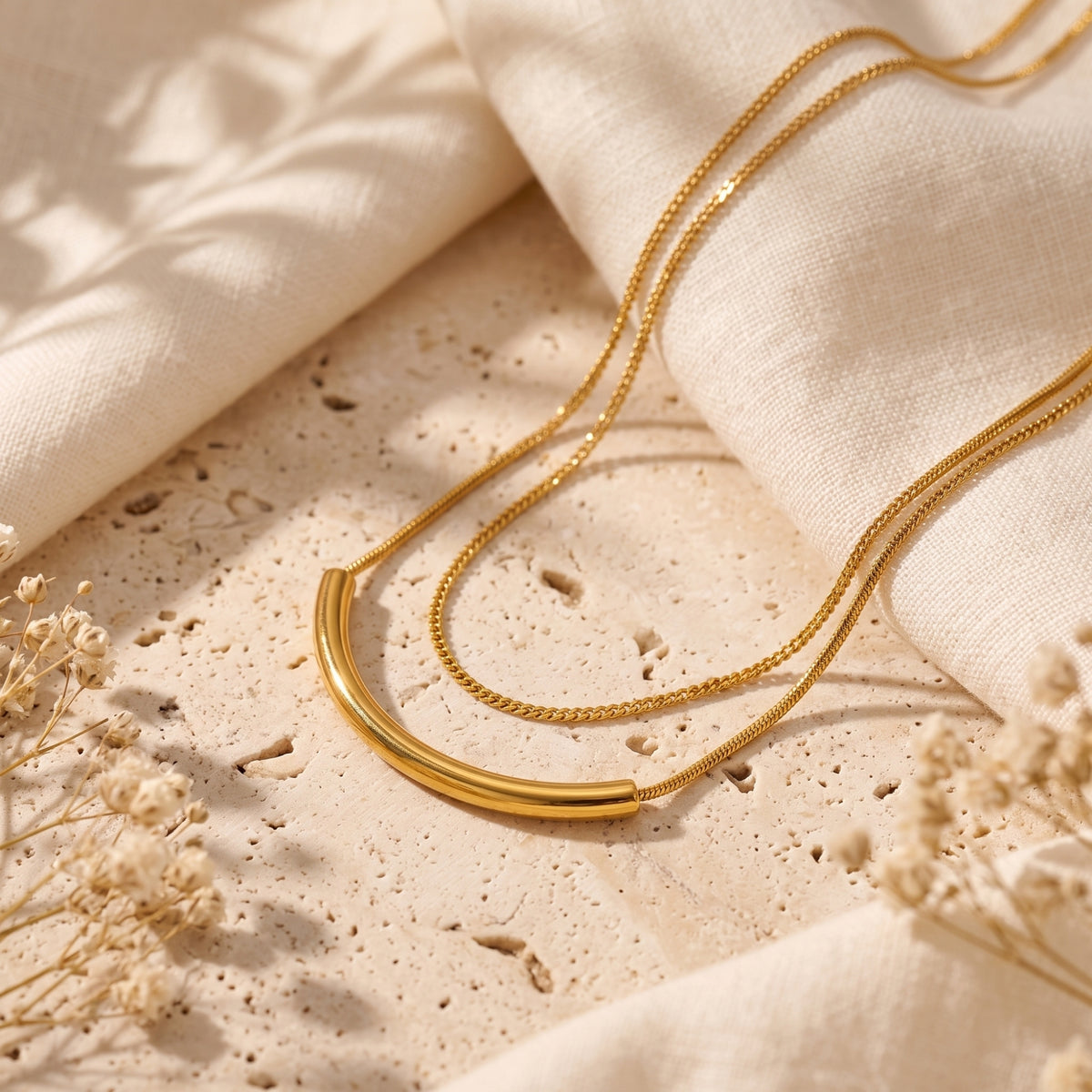 Gold-tone double-chain arc bar necklace resting on a textured beige stone slab with draped linen.