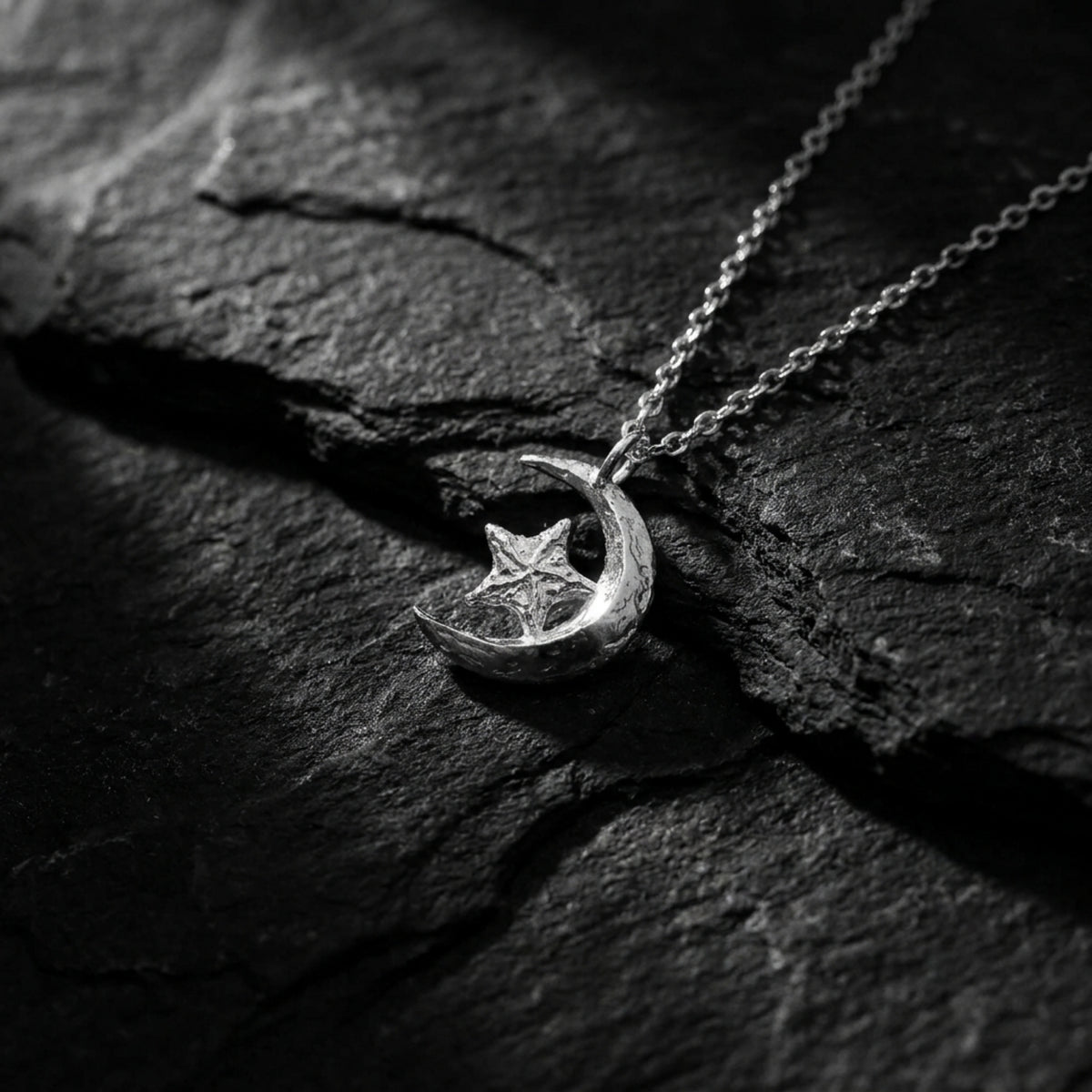 Silver crescent moon and star pendant resting on textured dark slate with its chain extending upward.