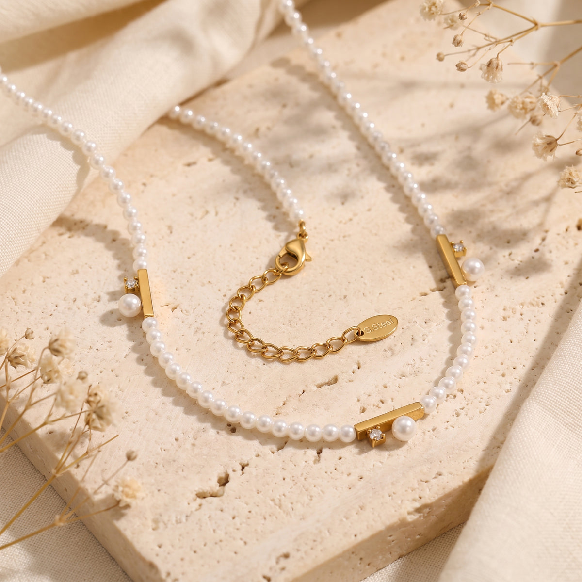 Close-up of the gold-tone pearl necklace showing its lobster clasp, extender chain, bar accents, and steel tag.