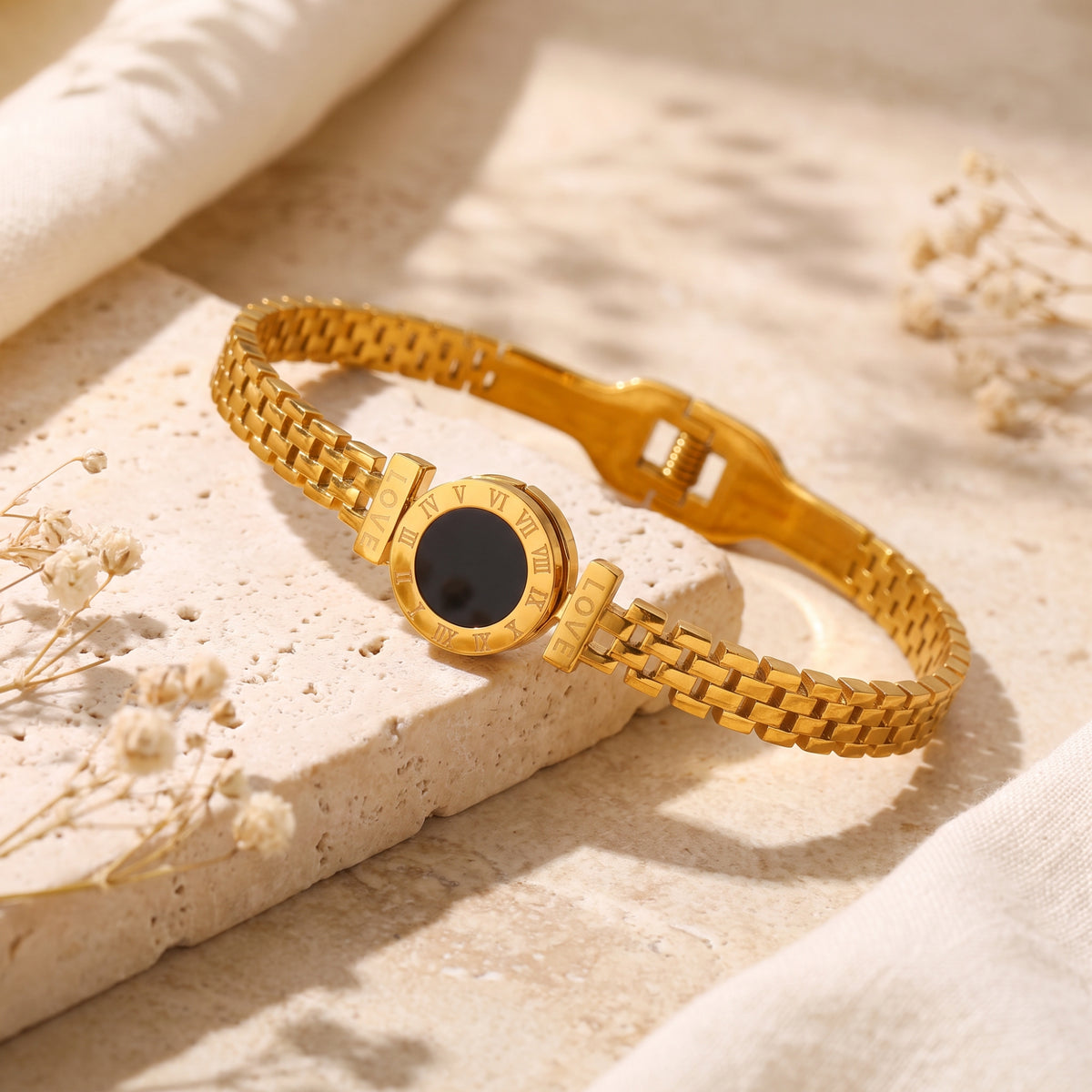 Angled view of a gold-tone link bracelet with a black Roman numeral medallion on a beige stone slab.