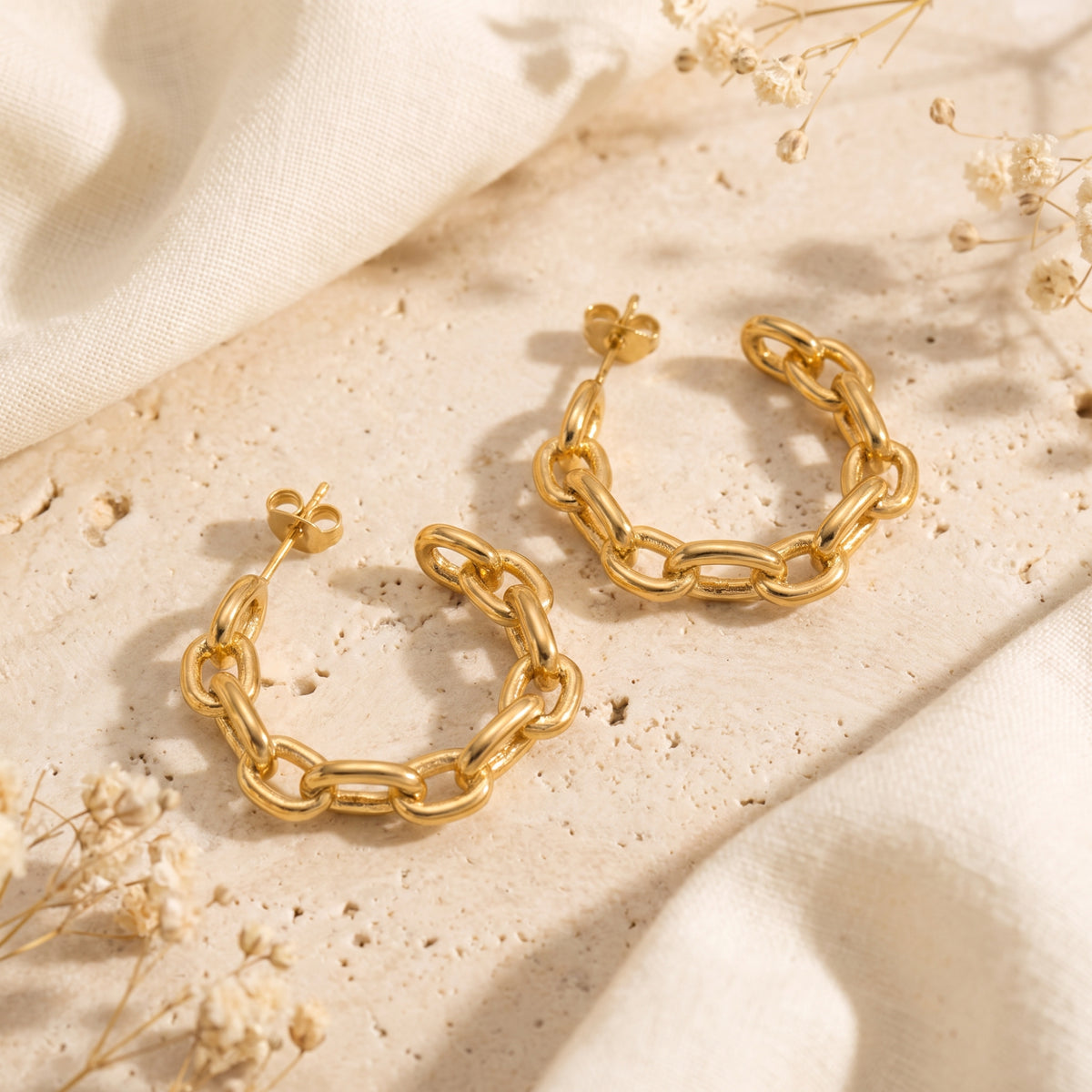 Pair of gold curb chain link hoop earrings lying flat on a textured stone surface beside white fabric.