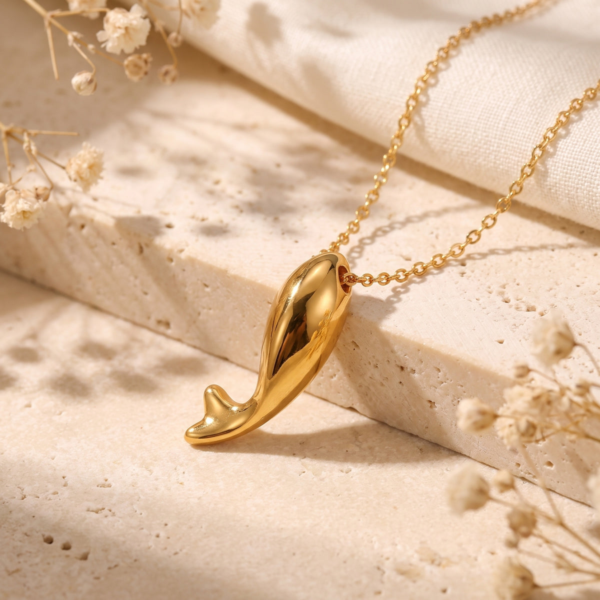 High-polish gold-tone dolphin tail pendant resting horizontally on a textured stone surface alongside its chain.